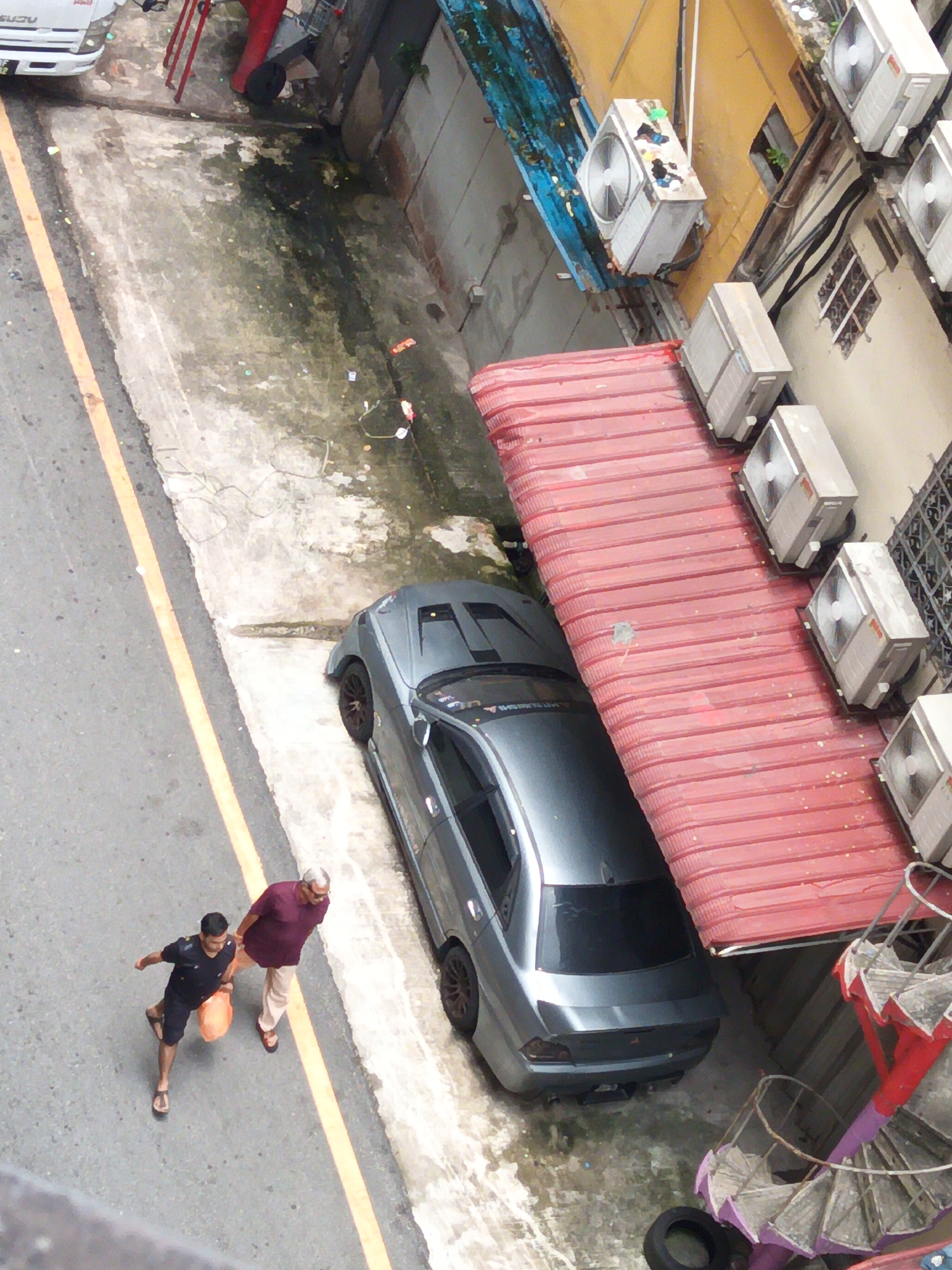 The image depicts an overhead view of an outdoor scene in Kuala Lumpur, Malaysia, likely a back alley or service area. In the foreground, two men are walking on a grey asphalt road marked with a yellow line. The man on the left wears a dark blue t-shirt and dark shorts and carries a bright orange bag. The man on the right wears a maroon polo shirt and light-colored pants, and appears to be walking alongside the other.
A silver four-door sedan is parked to the right of the men, partially covering a red corrugated metal structure that appears to be a garage door or awning. Several window air conditioning units are visible mounted on the exterior of a yellow building to the right and behind the red structure. A spiral staircase, painted red and purple with white steps, is situated to the far right of the image, adjacent to the building. The ground around the parked car and the red structure is a rough, concrete surface with patches of dampness and debris. In the upper left corner of the image, a white truck with the brand name "ISUZU" visible is partially captured.