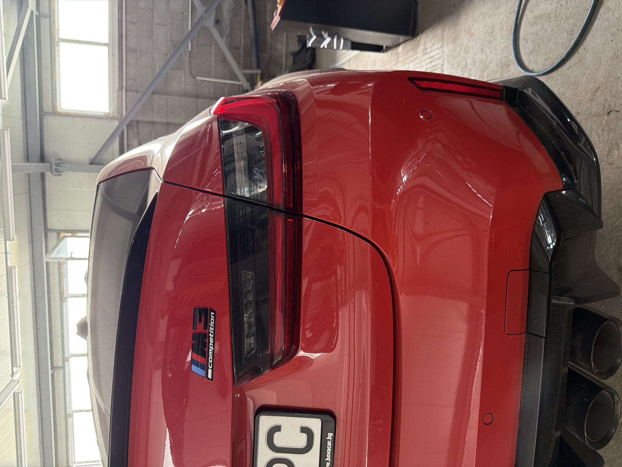 The image shows the rear-left quarter panel of a red BMW M3 Competition sedan parked indoors. The car has a black spoiler and black quad exhaust tips. A license plate reading "PC" and the website "www.bovacar.bg" is partially visible on the left side of the rear. The background consists of a concrete wall, industrial lighting fixtures, and large windows, suggesting a workshop or garage environment. The overall scene is well-lit.