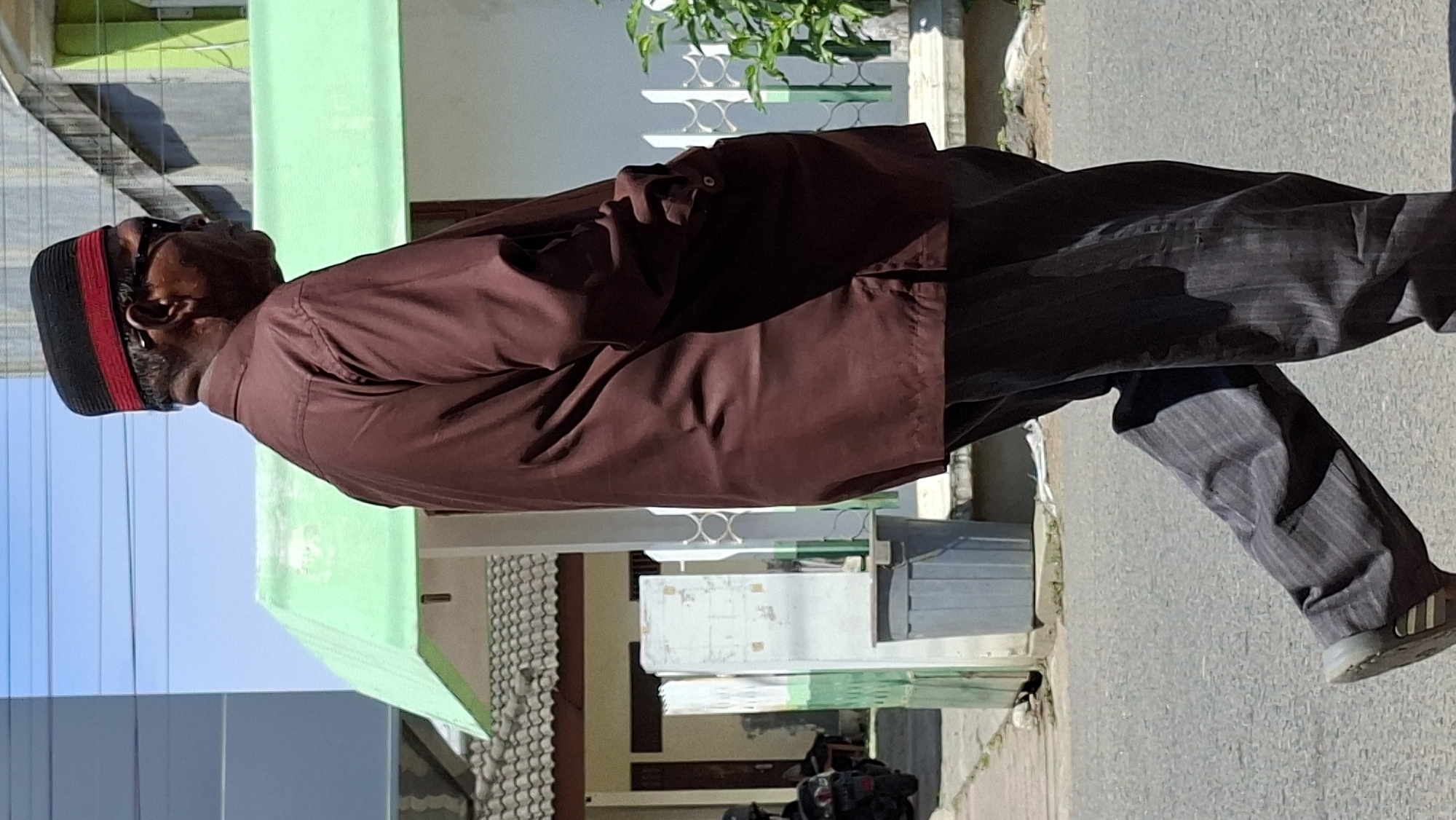 An elderly Indonesian man wearing a brown long-sleeved shirt, dark pinstripe pants, and a black and red kufi hat walks on a paved road in Lhokseumawe, Indonesia. The man is viewed from the side, walking from left to right. Behind him, a light green building with a tiled roof and white ornate fences are visible. A grey trash bin and a signpost are on the sidewalk. The sky is bright blue with scattered clouds. A motorcycle is parked to the left of the scene.