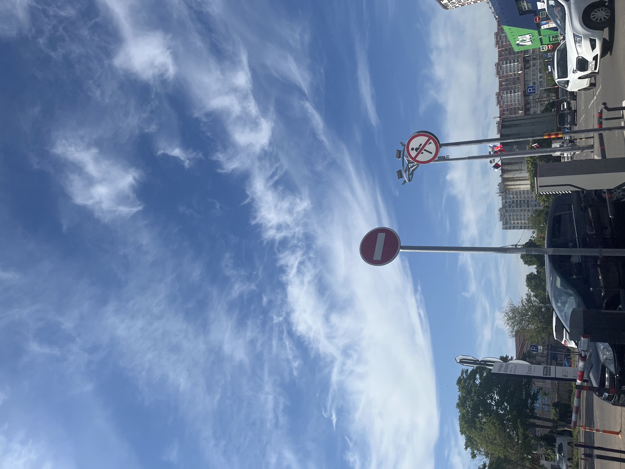 This is a vertical, outdoor, daytime photograph taken in Krasnodar, Russia. The scene captures a street with several cars and buildings under a bright blue sky with wispy white clouds.
In the foreground, a black car is parked on the left, partially obscuring a metal barrier. To the right of the black car, a white car is visible, with a portion of another white car further to the right. A parking sign with a blue "P" is seen in the background.
Two traffic signs are prominent. The one on the left is a circular red sign with a white horizontal bar, indicating "No Entry." To its right, mounted on a separate pole, is another circular red sign with a diagonal red bar across a black silhouette of a person walking, signifying "No Pedestrians." Both signs are mounted on metal poles.
Behind the traffic signs, a tall, multi-story building complex with numerous windows is visible, stretching across the mid-ground. The building appears to be residential. To the left of the building, a cluster of green trees stands. Several smaller buildings, some with colorful facades, are also present in the background.
The sky occupies the upper half of the image and is a vibrant blue, streaked with thin, feathery white clouds. The overall impression is that of a sunny day in an urban environment.