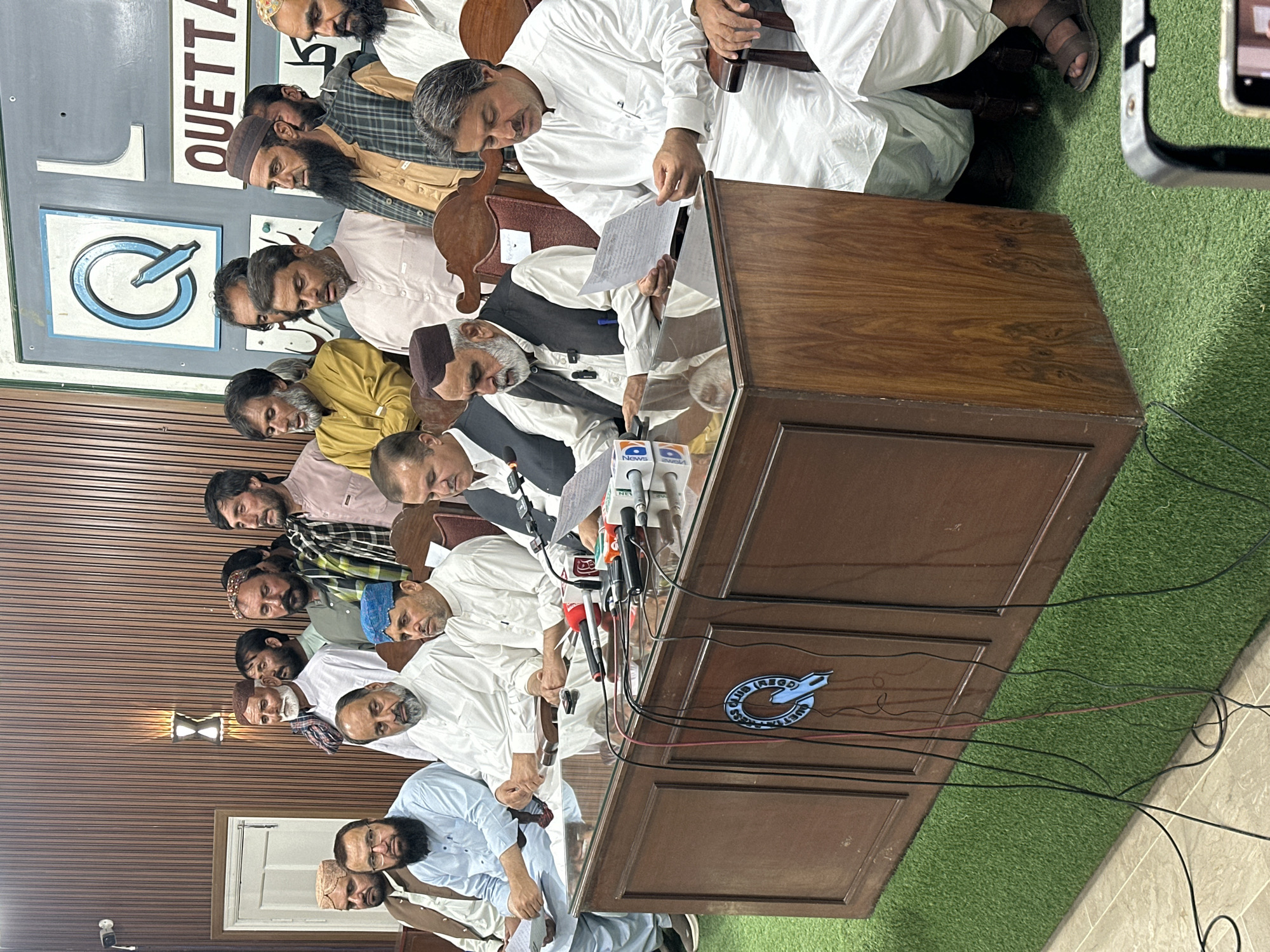 The image shows a group of approximately fifteen men seated at a large, dark brown wooden table. The men are of various ages and appear to be of South Asian or Middle Eastern descent. Many of them are wearing traditional clothing, including white robes and head coverings. Several microphones are on the table, suggesting a press conference or formal announcement. In the background, a sign with the word "QUETTA" is visible, indicating the location is Quetta City Tehsil, Pakistan. The setting appears to be indoors, possibly a meeting room or conference hall, based on the wall and the artificial grass rug in the background. The men are looking down, some holding and reading papers. Overall, the scene conveys a sense of seriousness and formality.