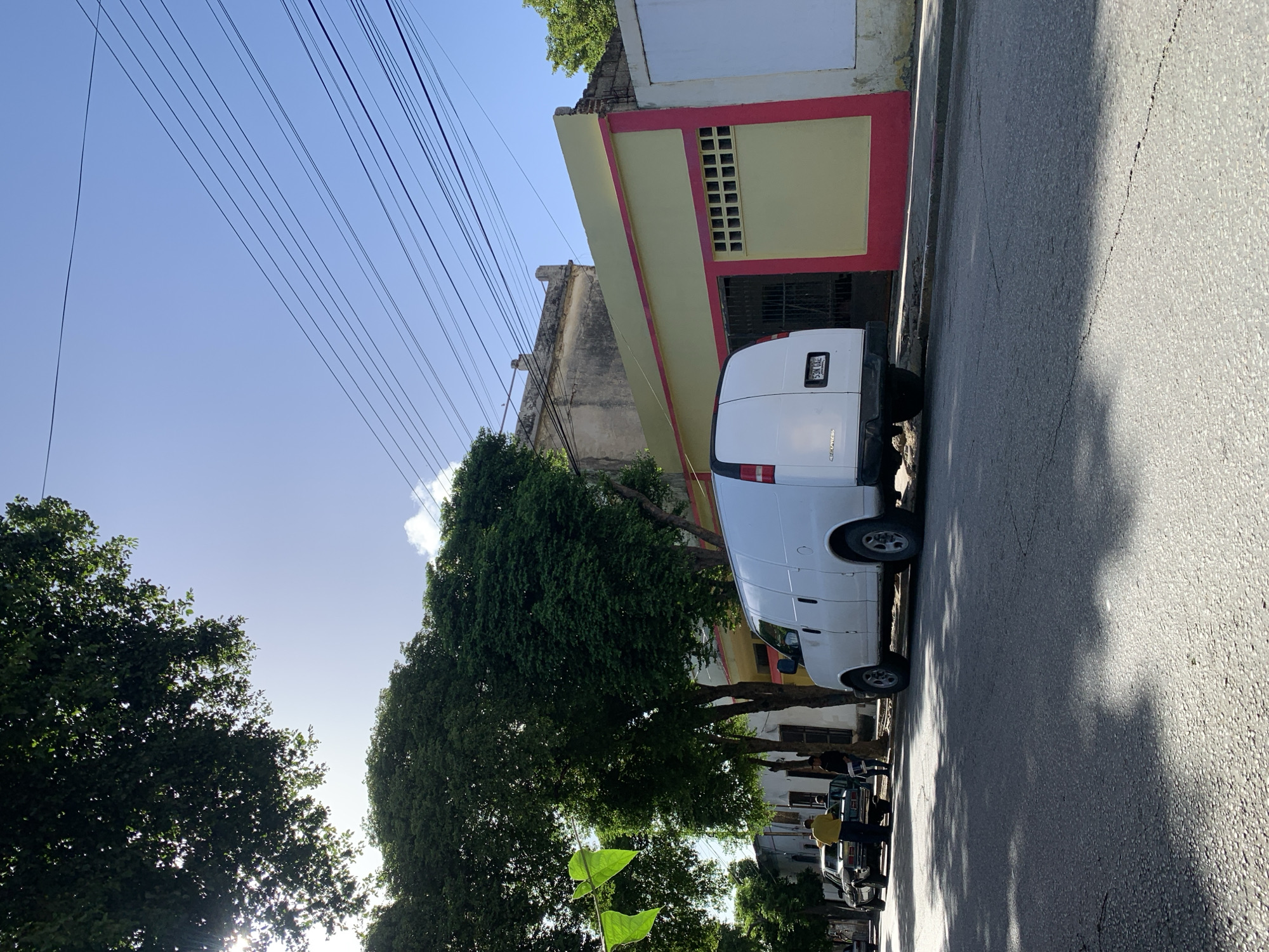 A white van is parked on the right side of a street in Santa Ana, Venezuela. The van faces left and is slightly angled towards the viewer. The street is paved and has some cracks. Trees with dense green foliage line the left side of the street, casting dappled shadows on the road. Beyond the trees and the van, a building with a yellow facade and a red trim is visible. The building has a gated entrance and some square openings. Power lines are strung across a clear blue sky. There are two cars parked further down the street on the left, and a few people are standing near them. One person is wearing a yellow shirt and appears to be talking to someone next to a dark-colored car. Another person in a dark shirt stands near the front of that car. The sun is shining, creating bright light and distinct shadows.