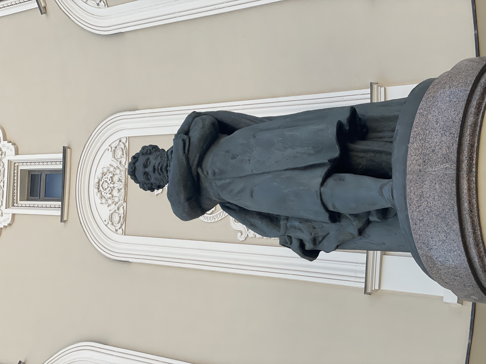 A low-angle, full-length photograph depicts a statue of a man in dark colors, positioned in front of a building facade. The statue stands on a circular base with a textured gray-and-black pattern. The man is dressed in a long coat, and his head is turned to the left. The building behind the statue is beige with white architectural details, including decorative molding around a window and a white, ornate frame behind the statue. The photograph was taken in the city of Kazan, Russia.