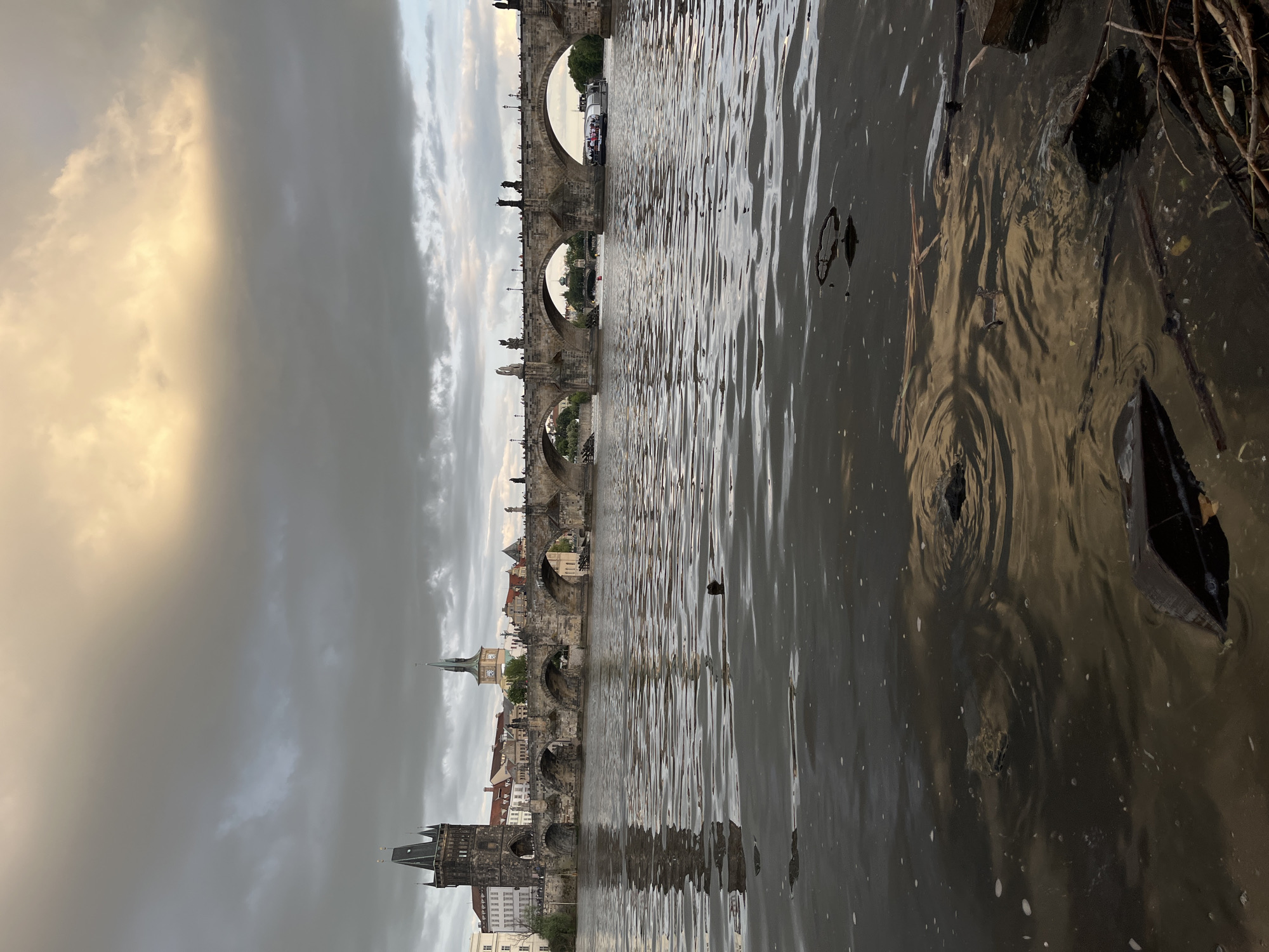 The image captures a serene view of a wide river flowing past a historic city under an overcast sky. The dark, rippled surface of the water dominates the foreground and midground, reflecting the muted light from above. Scattered debris, including branches and leaves, floats near the right edge of the frame.
A majestic multi-arched stone bridge, adorned with numerous statues along its balustrade, spans the river in the midground. Several arches of the bridge are visible, with reflections of the distant cityscape appearing in the water beneath them. A white passenger boat, carrying what seem to be people, is situated under one of the bridge's arches, suggesting ongoing river activity.
Beyond the bridge, a distinctive skyline of historic European buildings unfolds. On the left, a prominent dark gothic tower with a pointed spire and archways stands out, identifiable as the Old Town Bridge Tower. Further along the riverbank, other old buildings and a lighter-colored spire with a visible clock face are discernible.
The sky above is heavily overcast with significant grey cloud cover. However, a noticeable break in the clouds on the left reveals a patch of soft, golden light, suggesting either sunrise or sunset, or a moment where the sun briefly pierces through an otherwise cloudy day. The overall lighting creates a subdued and somewhat moody atmosphere. No visible text is present in the image. The scene, with its iconic bridge and architectural features, strongly indicates the Vltava River and the Charles Bridge in Prague, Czech Republic.