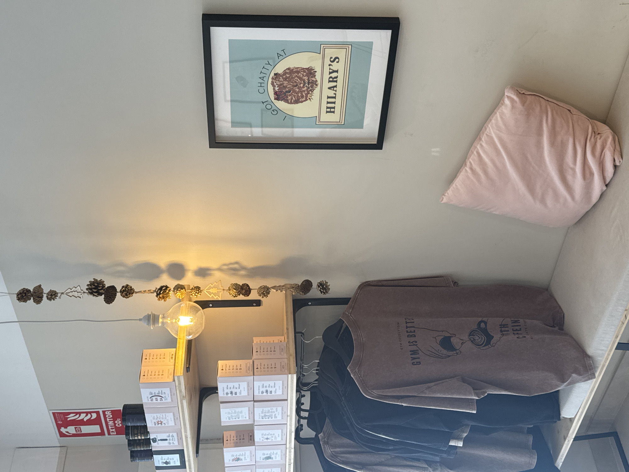 The image depicts an interior scene, likely a cafe or retail space, with a minimalist aesthetic. A light fixture made of pinecones and wooden tree ornaments hangs from the ceiling, casting a warm glow. Below, shelves display boxes of various products. To the left, a red fire extinguisher sign is mounted on the wall. A rack of t-shirts is visible, with one prominently displaying the text "GYM IS BETTER" alongside an illustration of hands preparing a coffee. In the foreground, a light-colored bench is topped with a plush pink pillow. On the right wall, a framed artwork features a stylized illustration of a cat with the text "I GOT CHATTY AT HILARY'S." The overall environment suggests a cozy and curated space, possibly in the city of Murcia, Spain, given the fire extinguisher sign in Spanish. There are no people or active interactions depicted in the scene.