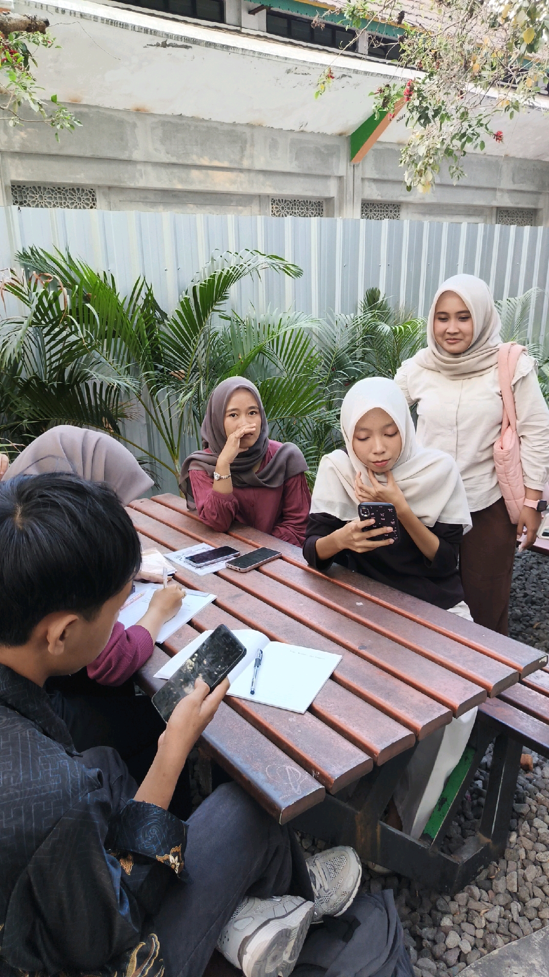 In Kartasura, Indonesia, a group of four young adults is gathered around a wooden picnic table outdoors. The scene appears to be a casual meeting or study session.
In the foreground, a young man with dark hair is seated, wearing a black patterned robe and grey pants. He is holding a smartphone and looking at it. To his left, a notebook and pen are visible on the table.
To the right of the young man, a young woman with a light-colored hijab is seated, wearing a black long-sleeved shirt. She is also looking at her smartphone, which she holds in her hands.
Behind her, another young woman wearing a grey hijab and a maroon long-sleeved shirt is seated. She has her hand near her face, with a thoughtful or pensive expression.
Standing behind the seated individuals is a young woman wearing a light-colored hijab and a beige button-up shirt, holding a pink bag over her shoulder. She is looking down and slightly to the right.
The environment suggests an outdoor seating area, possibly at a cafe or public space, with lush green foliage in the background and gravel on the ground. A grey corrugated fence and a concrete structure with decorative window grates are visible in the middle ground. The lighting suggests it is daytime, possibly late afternoon, given the shadows.