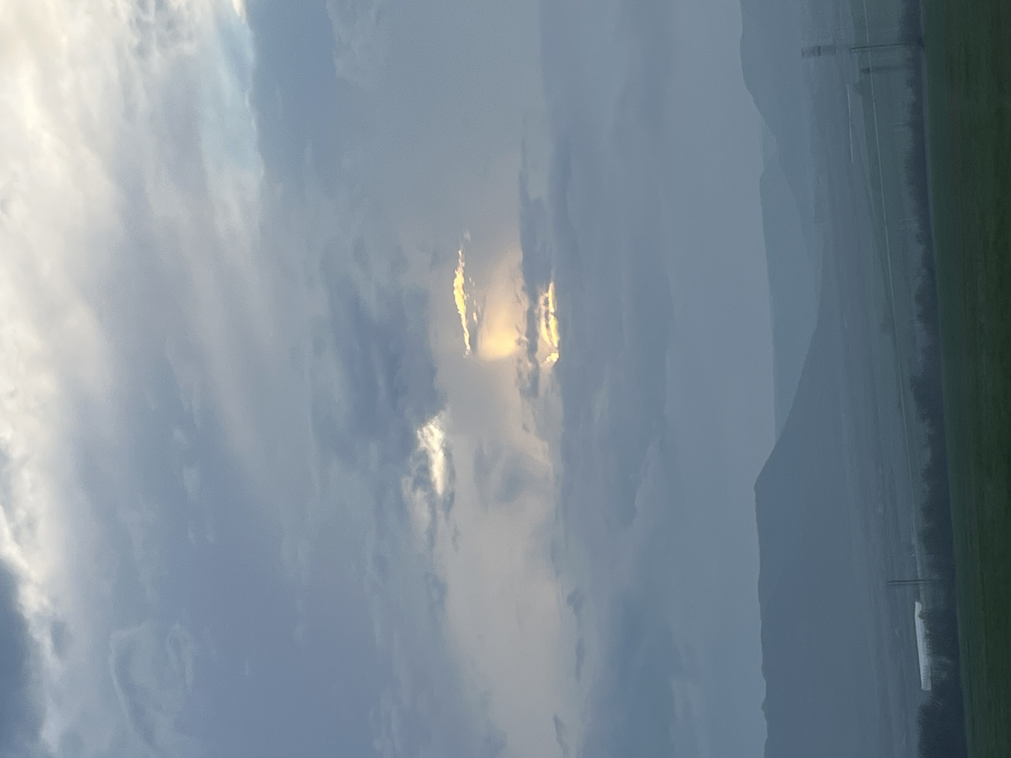 The image shows a landscape in Petrov Dol, Bulgaria. The sky is cloudy with a bright patch of sunlight breaking through the clouds. In the background, there is a mountain range obscured by haze. A green field with sparse trees and some infrastructure is visible in the foreground. The image is taken during the day.