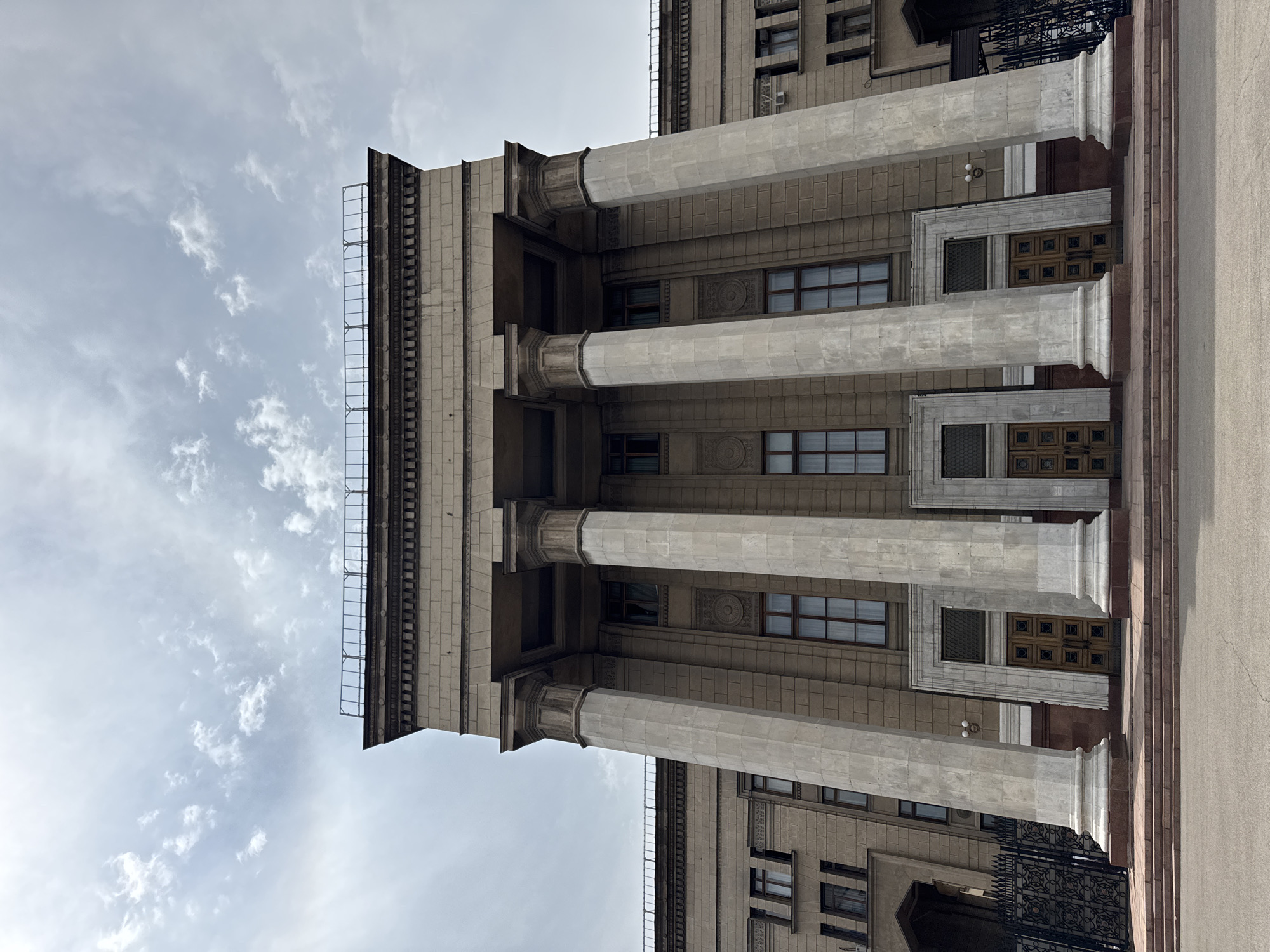 The image shows a low-angle view of a large, classical-style building, likely a government institution or university, with a grand entrance featuring a portico supported by four massive columns. The building is constructed of stone blocks and has multiple windows with dark frames. The sky above is overcast with scattered clouds. The scene is devoid of any people or vehicles. The architectural style and the presence of the columns suggest the building is a prominent landmark. This building is the Kazakh National University named after Al-Farabi, located in Almaty, Kazakhstan.