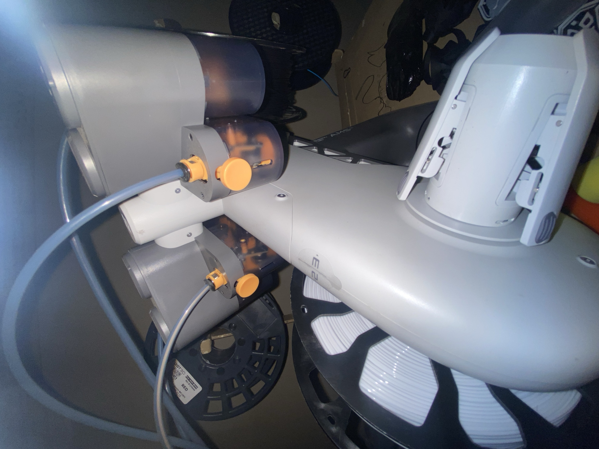 The image depicts a Bambu Lab AMS (Automatic Material System) for a 3D printer. The AMS unit is grey and white, with visible orange knobs on the feeder mechanisms. It holds multiple spools of filament, with one spool containing white filament and another containing black filament. A blue tube connects to one of the feeder mechanisms. The background is cluttered with miscellaneous items, including what appears to be a cardboard box and plastic bags. The image does not provide any information to determine the location in Buenos Aires, Argentina or any details about people, actions, or interactions.
