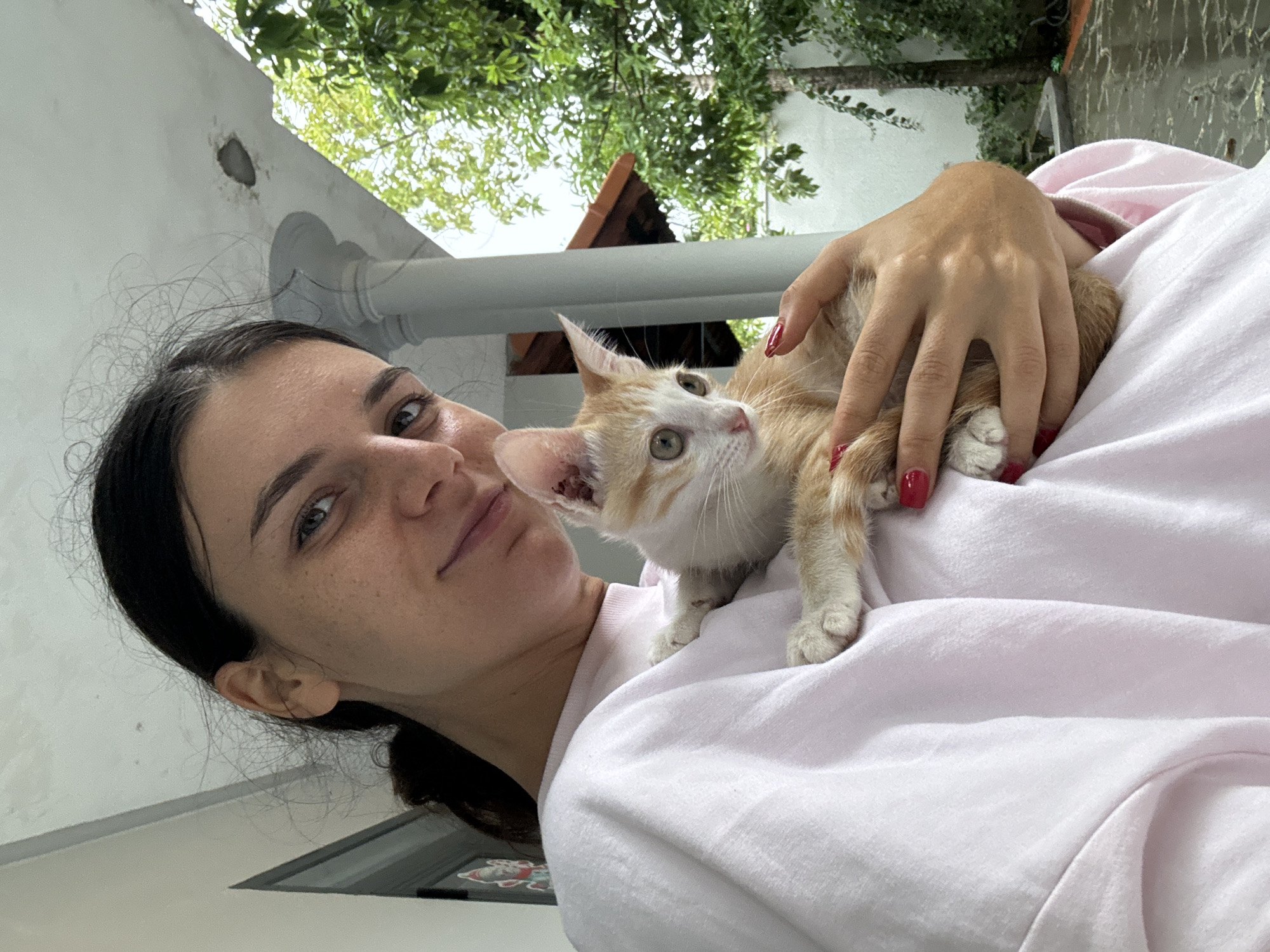 A young woman with dark hair tied back in a ponytail is holding a small, ginger and white kitten. She is wearing a light pink sweatshirt. The woman is looking at the camera with a slight smile. The kitten is looking to the side. They are on a covered porch or patio, with a white pillar and a roof visible in the background. Lush green trees and foliage are also visible in the background, suggesting an outdoor setting. The ground appears wet, indicating recent rain. The overall impression is one of a candid, affectionate moment between a person and their pet.
