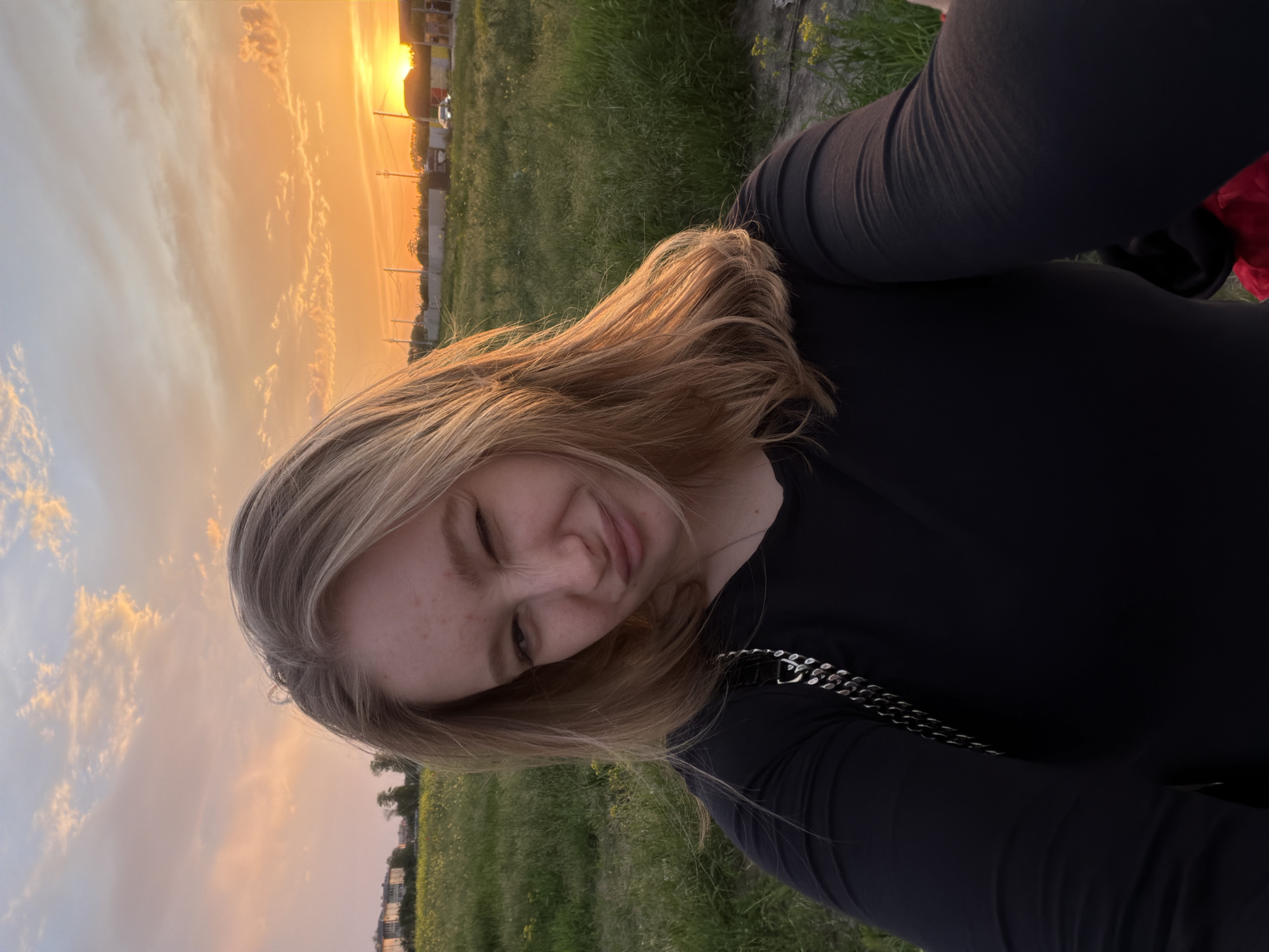 A young fair-skinned woman with shoulder-length blonde hair poses outdoors at dusk, near Krasnodar, Russia. She is wearing a dark long-sleeved shirt, and part of a silver chain is visible on her chest. She is smiling slightly with one eye closed as if winking. The sky is visible in the background with an orange glow from the setting sun and scattered clouds. Also visible is a field of grass and some residential buildings.