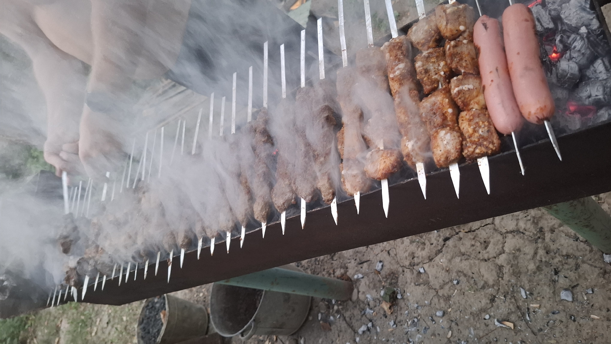 The image depicts an outdoor barbecue scene with numerous skewers of meat and two sausages cooking over hot coals. The scene is dominated by smoke rising from the grill, obscuring some of the details. Two individuals are partially visible in the upper right quadrant, with their hands engaged in managing the skewers. The barbecue grill itself is a long, dark metal structure, supported by green legs, positioned on uneven, dry, cracked ground. To the left of the grill, two metal buckets are visible, one appearing to contain charcoal. The overall environment suggests a casual outdoor gathering. The location is identified as Shubaragash, Kazakhstan.