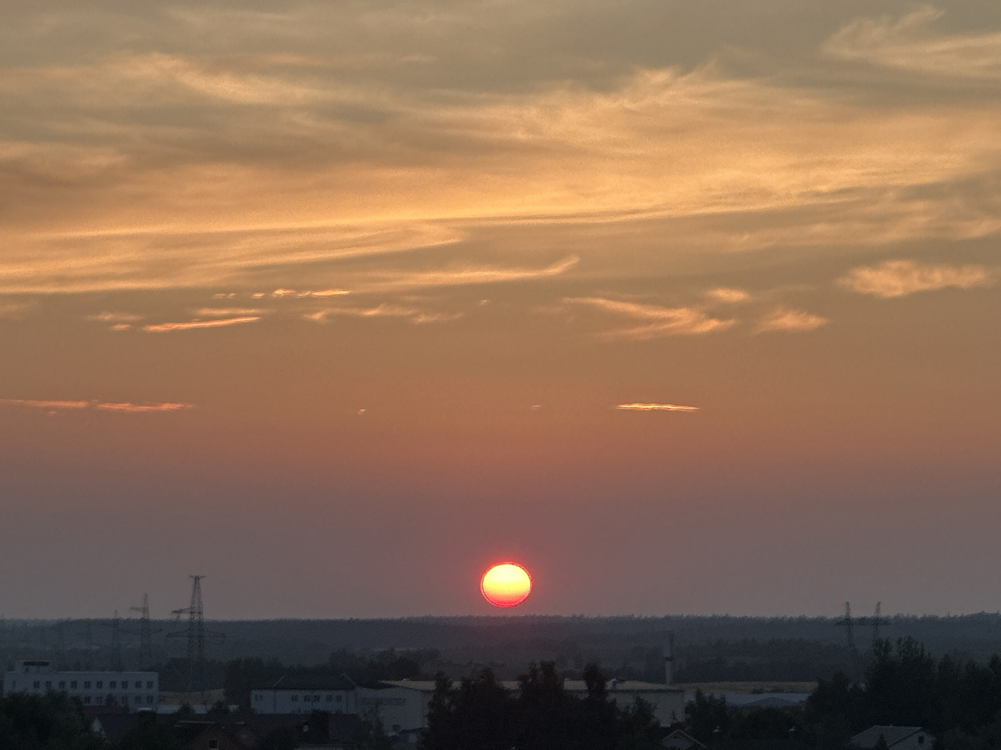 The image captures a sunset over a semi-urban area, likely on the outskirts of Шчомысліцкі сельскі Савет, Belarus. The sky is dominated by warm hues of orange and yellow, with wispy clouds stretching across the frame. A bright, orange-red sun is visible near the horizon, partially obscured by a dark, treed landscape that forms the foreground. Silhouetted against the sky are industrial structures, including tall power line towers and buildings, indicating a developed area. The overall impression is one of natural beauty meeting human infrastructure during the transition from day to night.