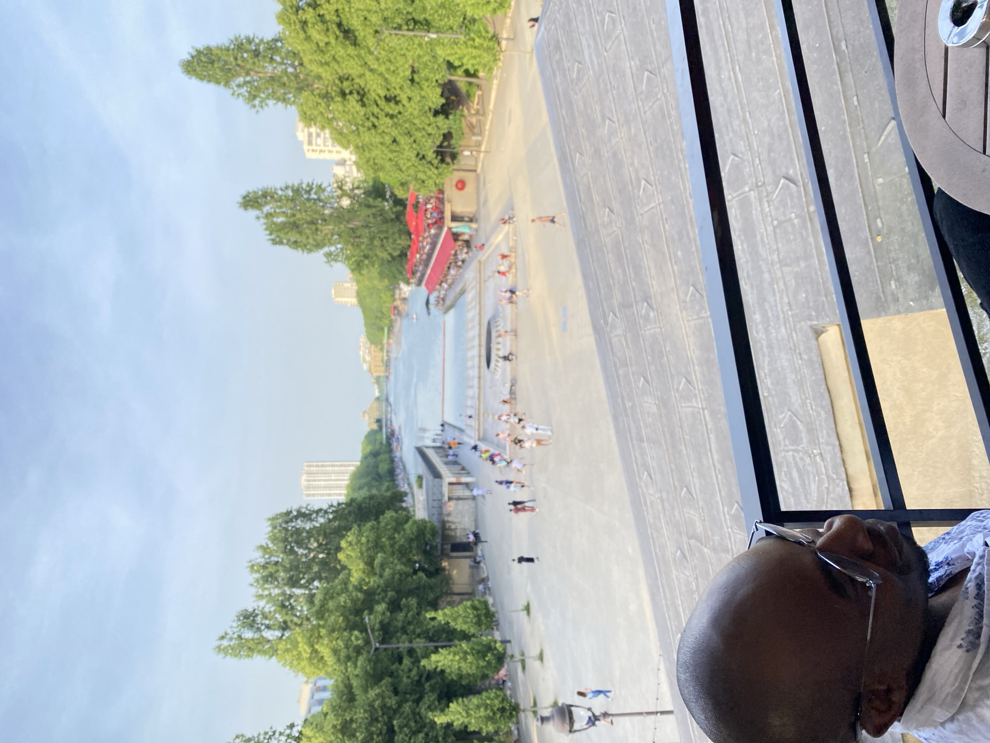 The image shows an elevated view of the Bassin de la Villette in Paris, France. People are visible along the quays and near the water. Trees line the edges of the basin, and buildings are visible in the background. A portion of a person's head and glasses is in the foreground on the right, and part of what appears to be a metal railing or structure is also visible.