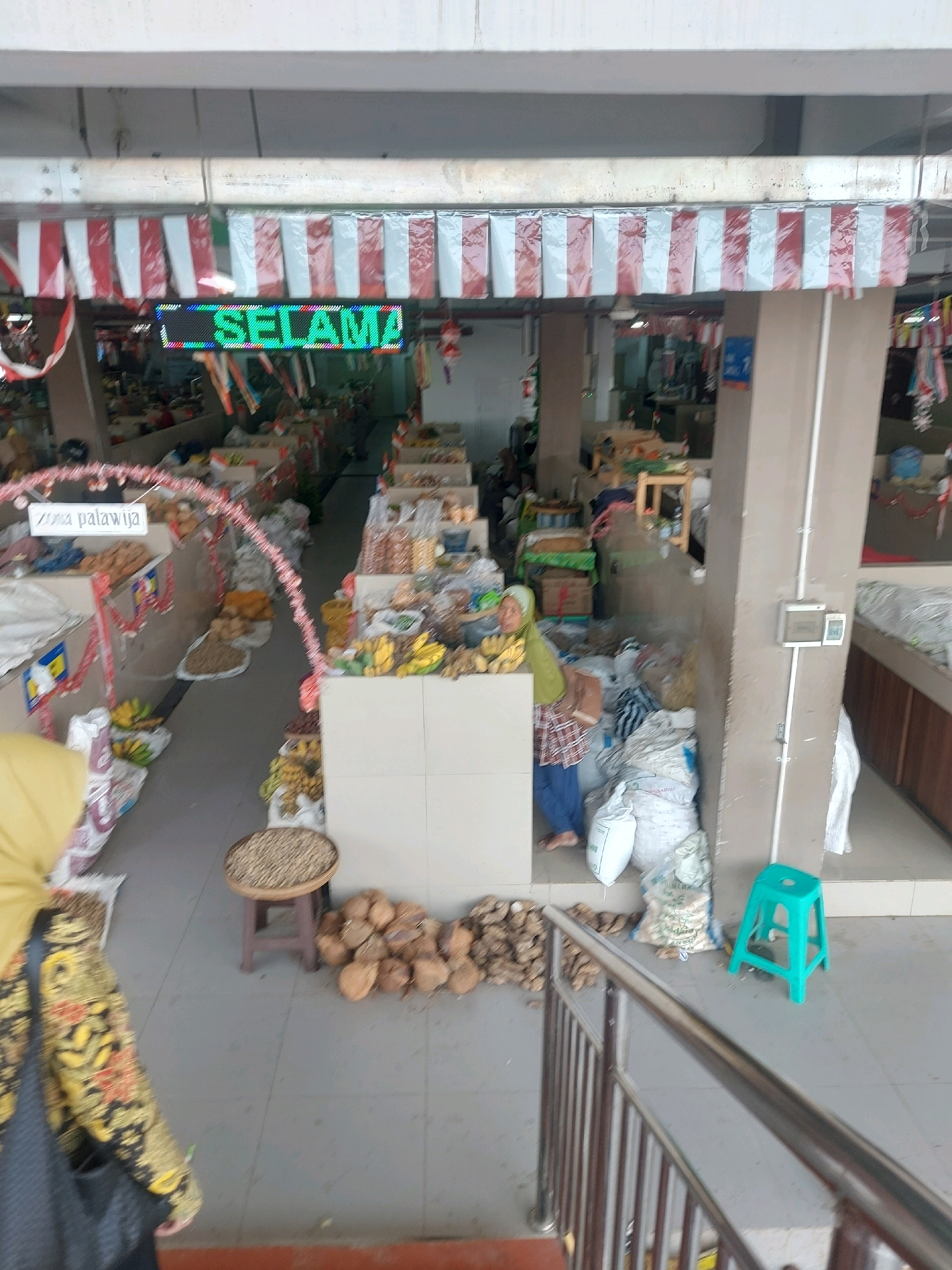 The image shows a market stall in Godean, Indonesia, with a woman in a headscarf and plaid apron standing behind a display of goods. In the foreground, a set of metal stairs leads up to the market level. The stall is decorated with red and white Indonesian flags, and a green LED sign displays the word "SELAMAT". Various produce, including bananas, coconuts, and what appear to be dried goods, are arranged on the floor and in baskets. Several sacks of goods are also visible, along with a small, teal-colored stool. The market appears to be an open-air or covered structure with concrete flooring and pillars. The lighting suggests it is daytime.