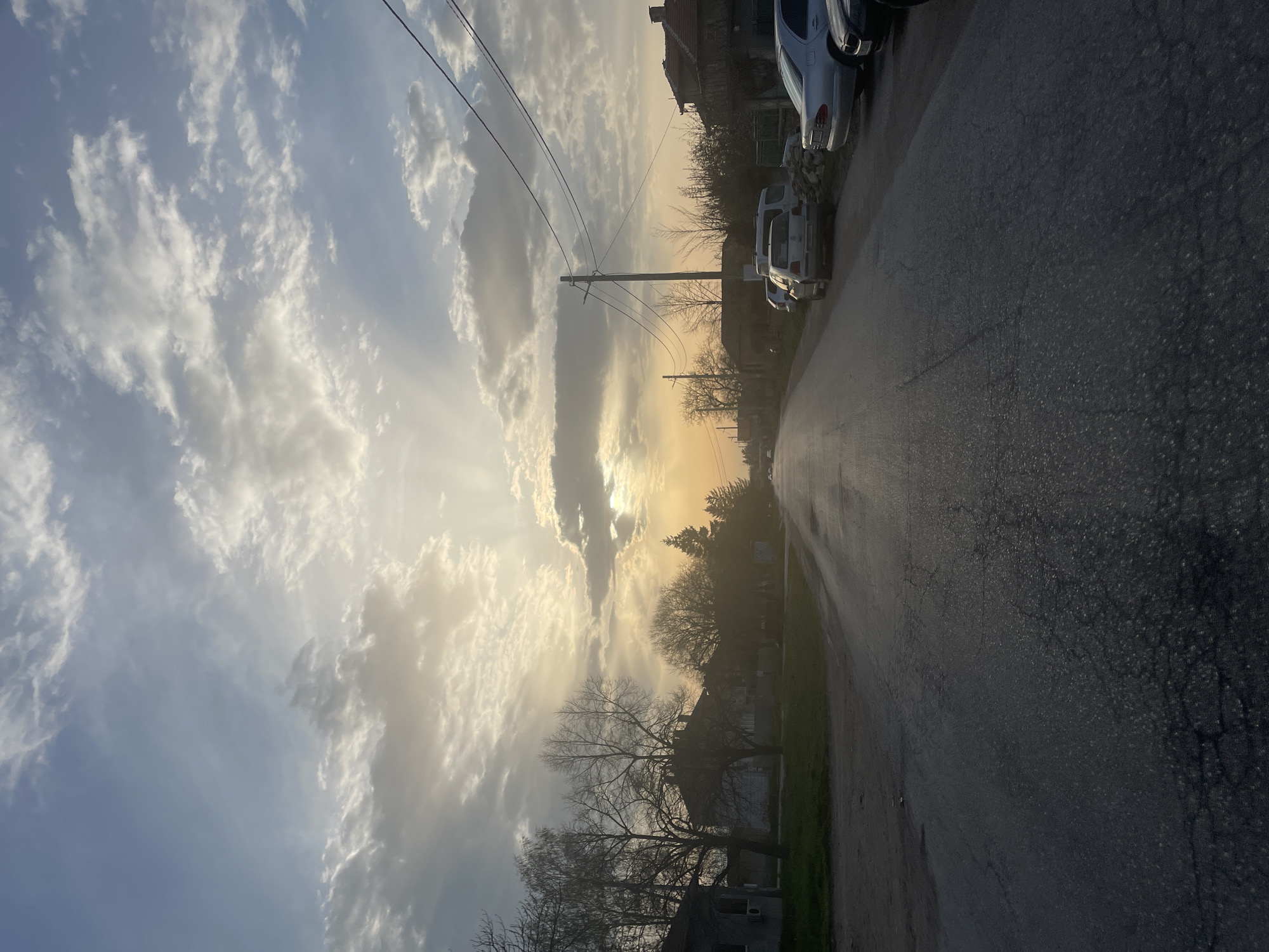 A medium-shot landscape photograph shows a street in Aprilovo, Bulgaria, during the day. The street runs diagonally from the lower left corner to the upper right corner. The sky is a mix of blue and white clouds. The sun is shining through the clouds, creating a glow. A power pole is visible, with wires strung across the sky. The street is wet and reflects the sky. There are cars parked on the right side of the street. Buildings and trees line the left side of the street. A few bare trees are in the foreground. The overall scene depicts a moment of sunlight and reflections on a street in Aprilovo.