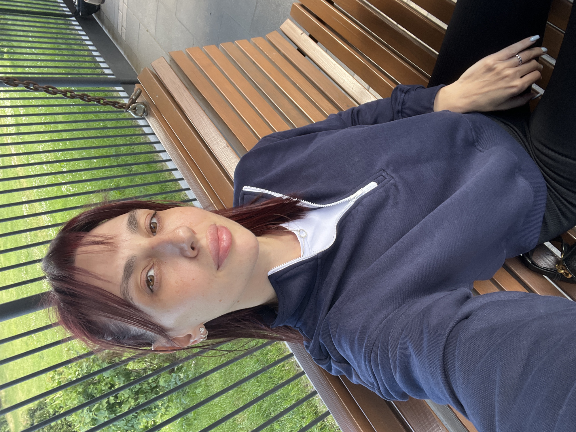 A medium shot, taken outdoors, shows a young woman sitting on a wooden swing bench in Nizhny Novgorod, Russia. The woman has dark red hair with bangs and is wearing a dark blue zippered sweatshirt over a white t-shirt. She is looking directly at the camera and has light makeup on. She is wearing small hoop earrings and a ring on her right hand. The swing bench is made of light brown wooden slats. Behind the bench, a black metal fence runs horizontally, and beyond that, green grass and foliage are visible. The image appears to be a selfie taken with a mobile phone, with the woman's left arm and hand visible in the lower left corner of the frame. The lighting suggests it is daytime.