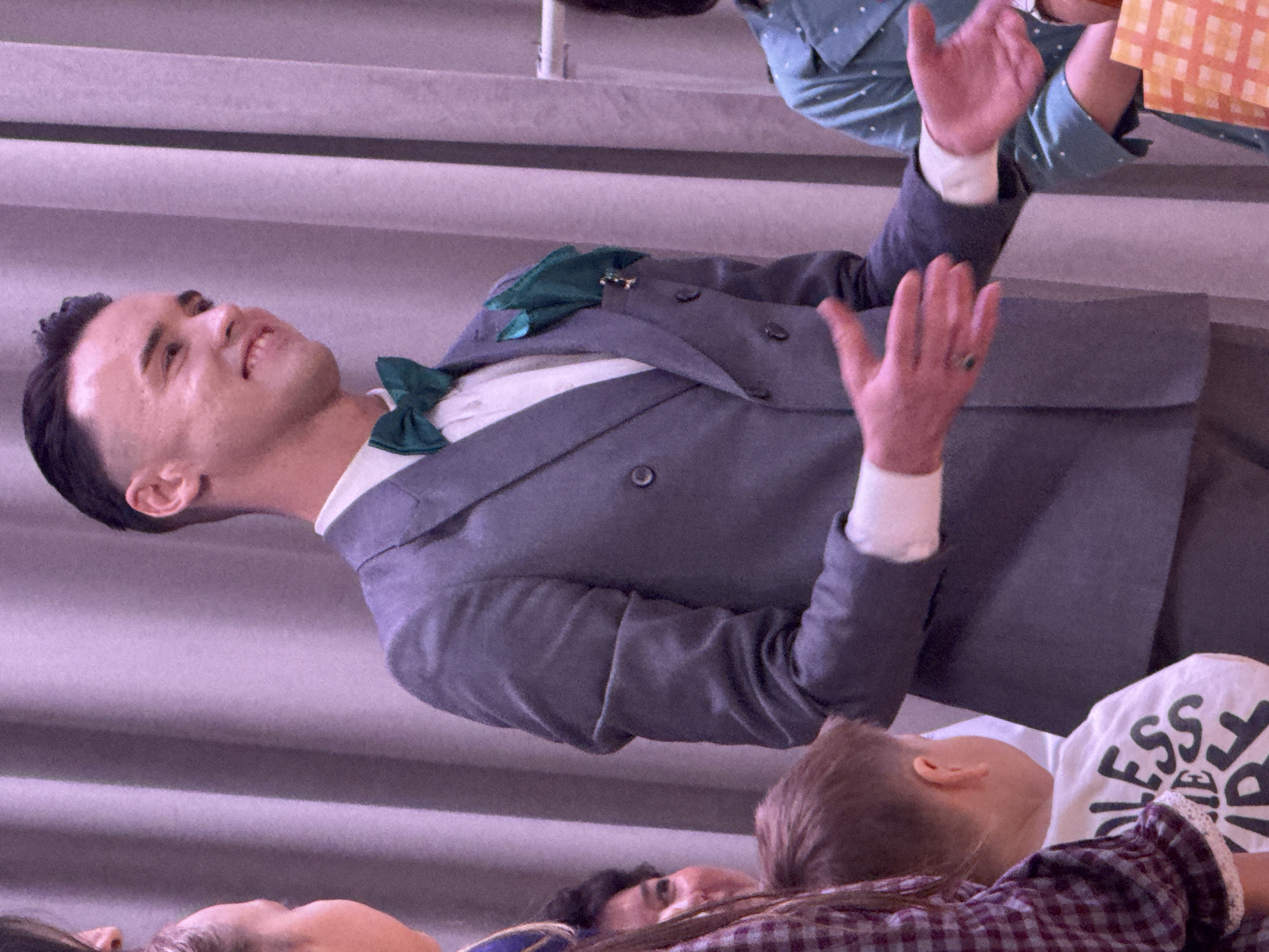A man in a grey double-breasted suit with a green bow tie and pocket square stands on a stage, his hands raised, seemingly engaged in a performance. He is surrounded by audience members, including children, whose faces and clothing are partially visible at the bottom of the frame. The stage is backed by a wall with horizontal lines. No information confirms or denies that the location of the image is Высокая Гора, Russia.
