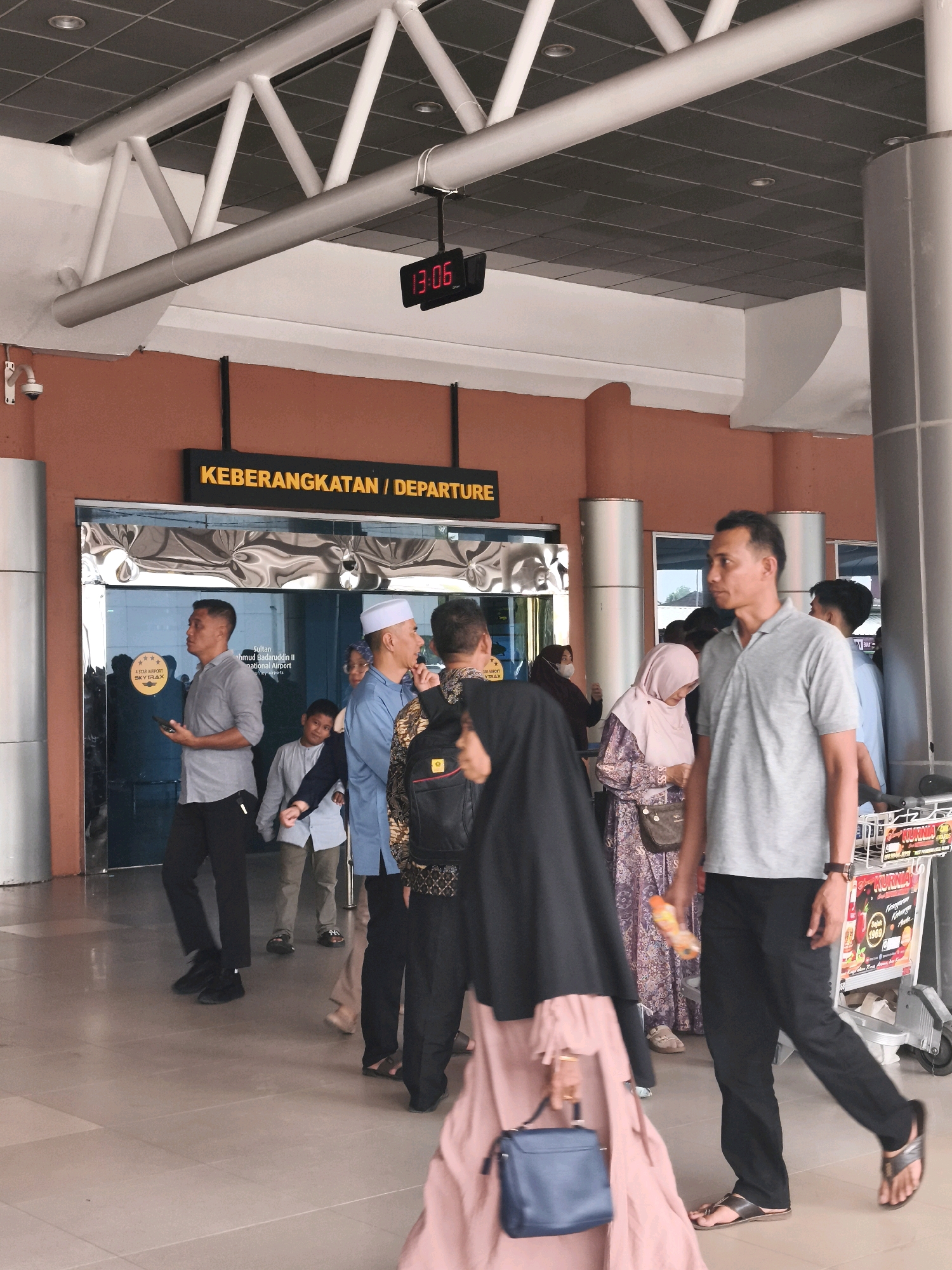 The image depicts the departure terminal of Sultan Mahmud Badaruddin II International Airport in Palembang, Indonesia. A digital clock on the ceiling displays "13:06". Several people, appearing to be travelers and airport staff, are visible in the scene. Some individuals are dressed in traditional attire, such as a man wearing a white peci cap and another in a batik patterned shirt. Others are in casual Western-style clothing, including t-shirts and trousers. Several people are carrying bags, and some are near a baggage cart. The architecture features large pillars, a metal truss ceiling, and an orange-colored wall with signage indicating "KEBERANGKATAN / DEPARTURE". The flooring is tiled. A security camera is mounted on the wall. The overall atmosphere suggests a busy airport environment.