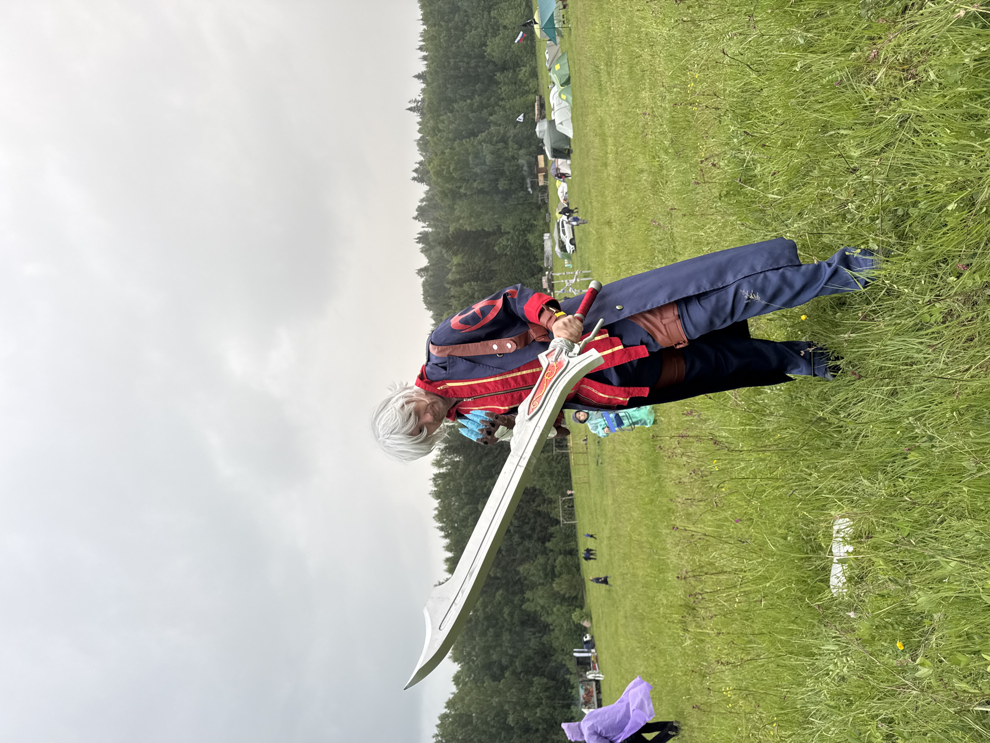This image depicts an outdoor event in a large grassy field located in Большая Кивара, Russia, under an overcast sky. The central figure is an adult individual with white, spiky hair, dressed in a navy blue long coat featuring red trim and a red circular emblem on the left arm, worn over a red zippered top. Brown leather-style straps are visible on their shoulder and waist. They wield a large, stylized sword with a silver blade and a red and gold hilt, held across their body. Their right hand wears a blue glove. The individual's gaze is directed downward and to their right.
In the mid-ground to the right, a temporary encampment consists of several tents in various colors (green, white, blue) and a parked white car. A Russian flag and another light-colored flag are displayed on poles nearby. Multiple other people are present in the field, including a person in a purple poncho on the far left and a child in a light blue outfit behind the main subject. The field is covered in green grass with scattered small yellow and purple flowers. A dense forest line forms the background. The sky is uniformly light grey and cloudy. White debris is present in the grass in the foreground.