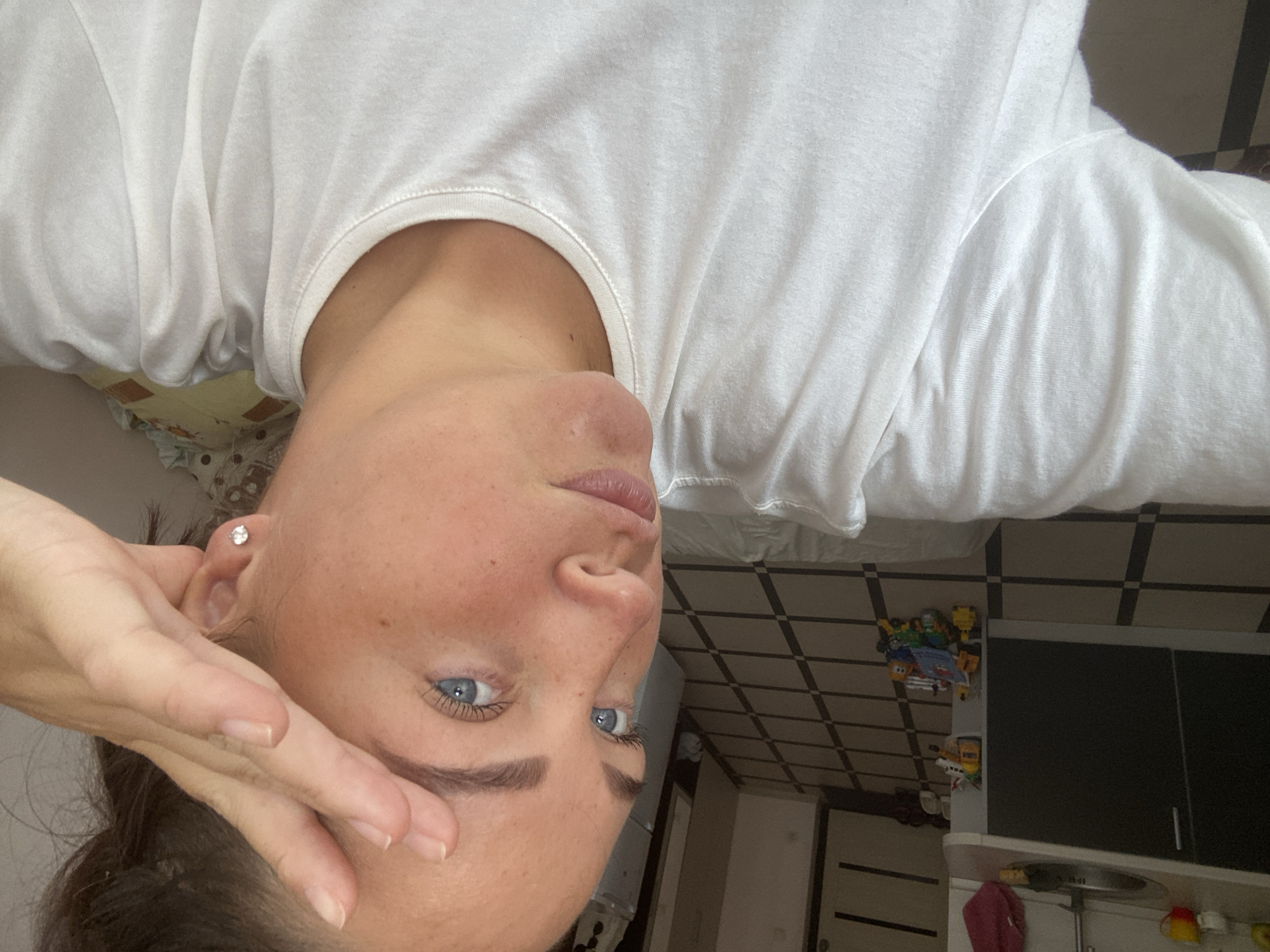 The image shows a close-up selfie of a woman with blue eyes and light skin with freckles. She is wearing a white t-shirt and has her hand partially covering her forehead. She is wearing a small diamond stud earring in her right ear. The background appears to be an indoor setting, possibly a kitchen or hallway, with tiled flooring and some toys scattered on the floor. There is no visual information in the image that can be used to determine the location within the city of Sochi, Russia.