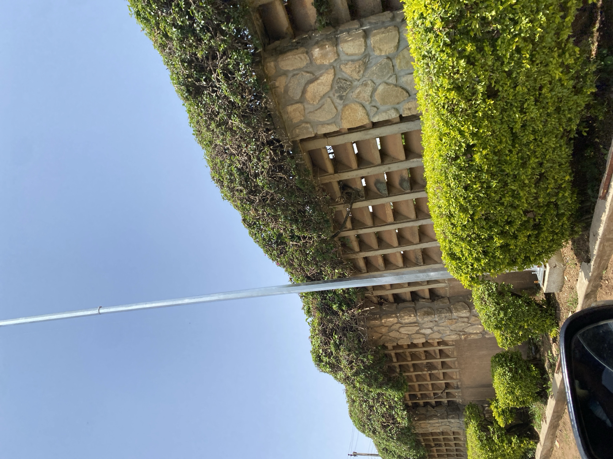 In Gura Top, Nigeria, a stone and concrete wall, partially covered in lush green foliage, stretches across the frame under a clear blue sky. The wall features decorative stone pillars and a lattice-like concrete section. In front of the wall, manicured green bushes form a dense hedge. A tall, metallic pole, likely a lamppost, stands vertically on the right side of the image. The foreground shows the edge of a road and a portion of a car's rearview mirror.