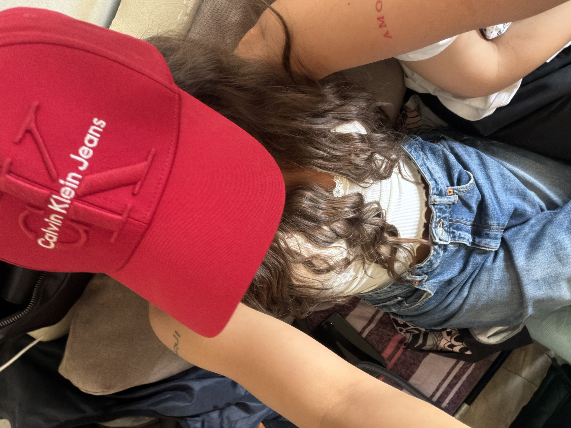 A person wearing a red Calvin Klein Jeans baseball cap, a white top, and blue denim jeans is lying down on a couch. The person has long, wavy brown hair. There are visible tattoos on their arms. The environment appears to be indoors, possibly a living room, with luggage or bags in the background. The image does not provide any specific visual cues that would allow for the identification of the location within Varna, Bulgaria.