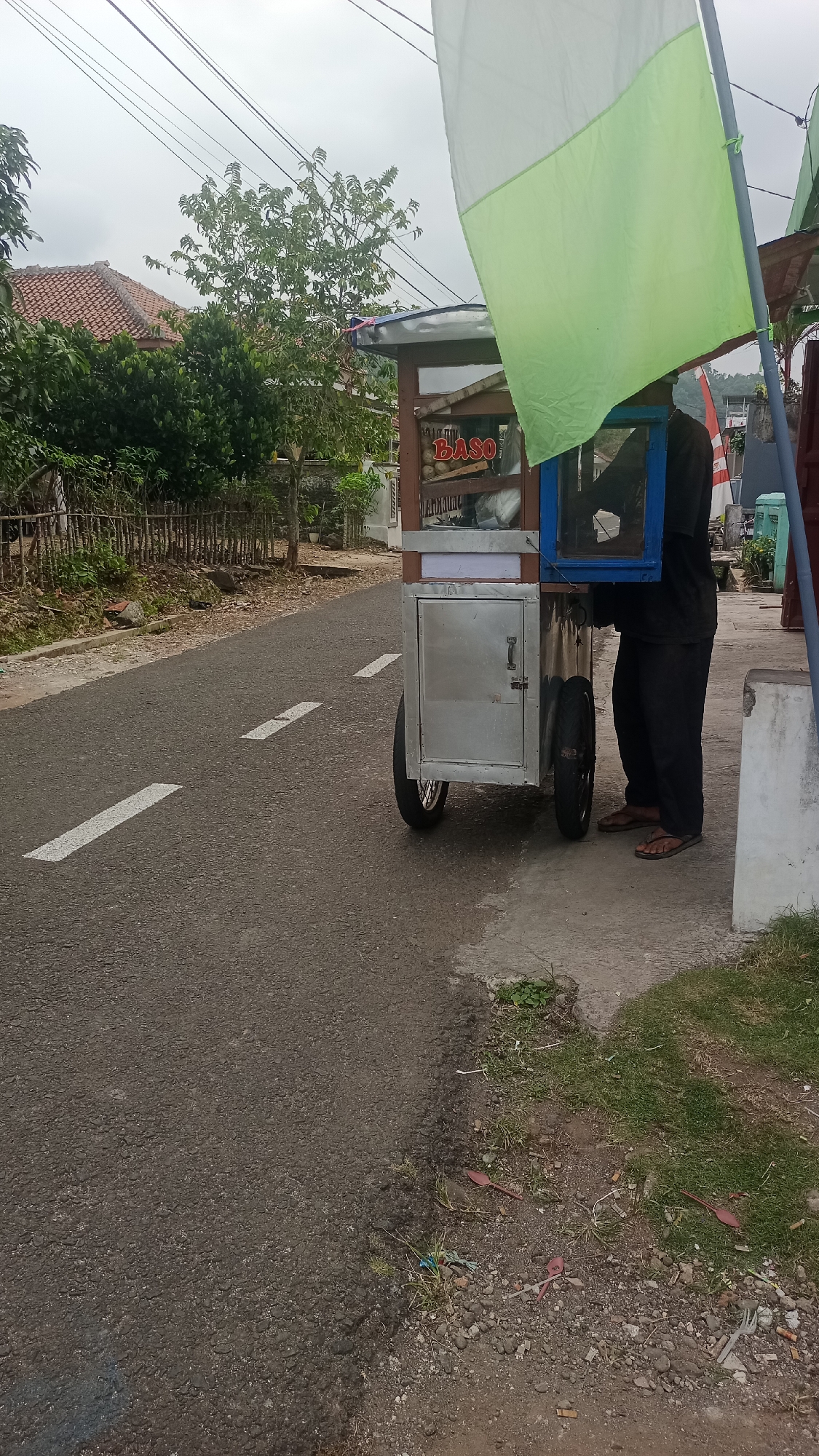 A street vendor is preparing food from a cart on the side of a road in Pakemitan, Indonesia. The street is paved with asphalt and has white lane markings. A cart with wheels is positioned on the right side of the road, with a person in dark clothing attending to it. The cart has a metal body and a glass display case. A green and white flag is attached to a pole next to the cart. The background shows trees, houses with red tile roofs, and power lines. The ground next to the road is covered in grass and dirt, with some scattered debris including plastic spoons and cigarette butts.