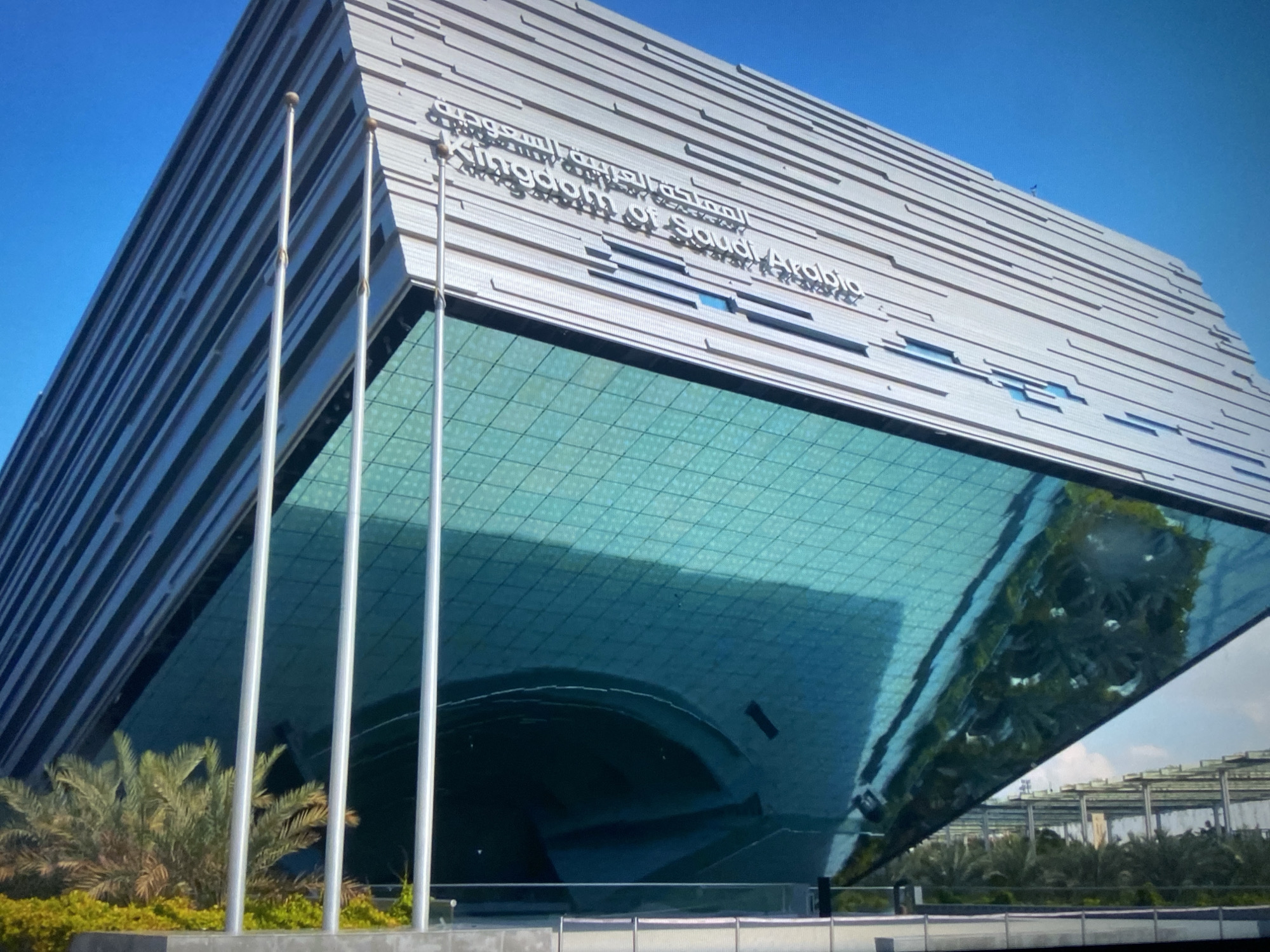 The image shows a modern building with a unique architectural design, identified by the text "Kingdom of Saudi Arabia" on its facade. The building features a rectangular upper section with horizontal linear patterns and a lower section constructed with reflective glass, creating a distorted reflection of the surrounding environment. Three flagpoles stand in front of the building, partially obscured by greenery. The scene suggests a clear day with a bright blue sky in the background. The location is possibly a large urban complex, based on the architectural style and surroundings.
