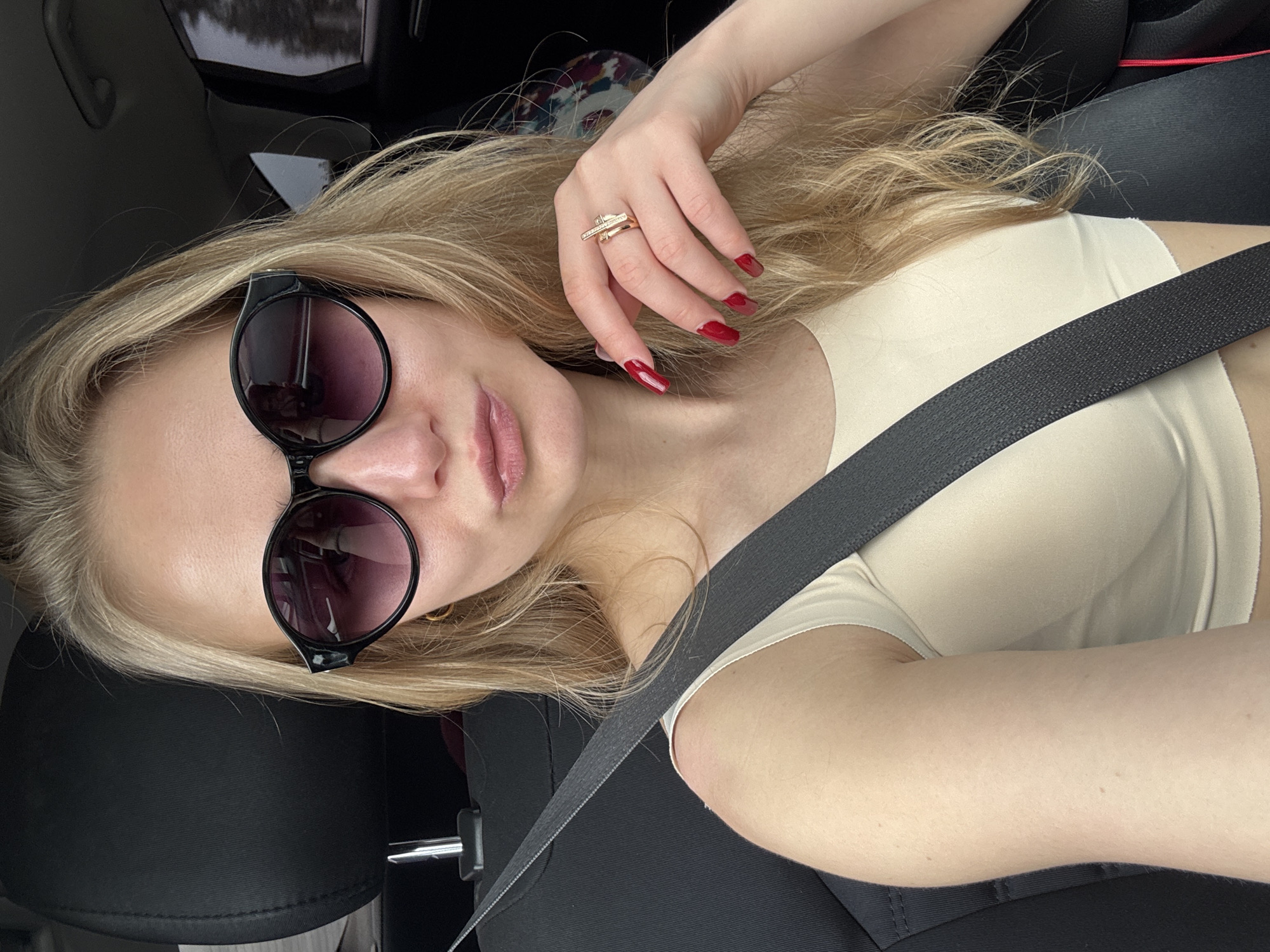 A woman with long blond hair and wearing sunglasses is sitting in a car. She is wearing a light beige top and has a seatbelt across her chest. Her right hand is near her head, and she has a gold ring on her ring finger. She has long red nails. The car seat and interior are black. The location is unknown.