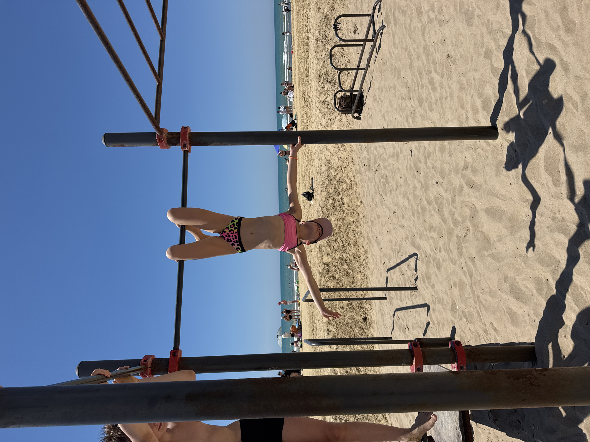 The image shows a young person performing a handstand on a bar at a beach fitness area. They are wearing a pink sports bra and leopard-print bikini bottoms. The background consists of a sandy beach with people in the distance, the ocean, and a clear blue sky. The sun is high in the sky, suggesting it is daytime. The scene is set at a beach, likely in a warm climate, with parallel bars and other fitness equipment visible. The overall mood appears to be one of active recreation and enjoyment of the outdoors. No text is visible in the image. The location is identified as Natukhaevskaya, Russia.