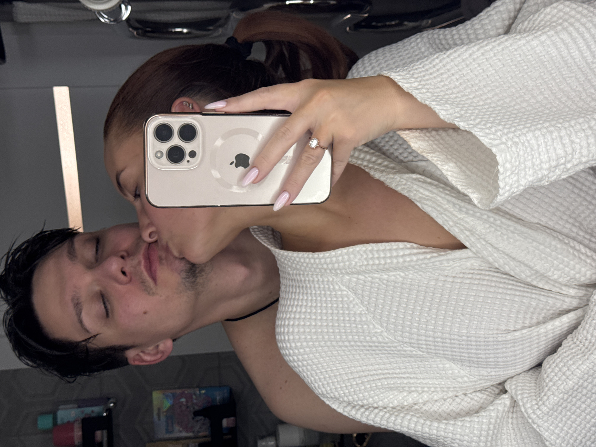 A couple is taking a selfie in a mirror. The woman is holding the phone, her face close to the mirror, while kissing the man, who has his eyes closed. The woman has reddish-brown hair pulled back in a ponytail and is wearing a white waffle-weave robe, which is partially visible. She is also wearing a ring on her left ring finger. The man has dark hair and is also possibly wearing a black cord necklace. The background shows a bathroom counter with various items, including a light fixture, and tiles on the wall. The location is potentially a private residence, but the image does not provide enough information to determine if it's in Chaikovsky, Russia.