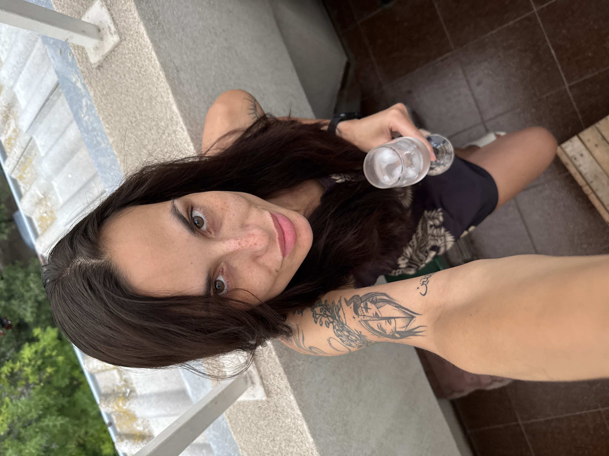 A woman with brown hair and freckles on her face is taking a selfie while holding a glass with ice inside. She is wearing a black dress with a floral pattern, and has tattoos on her left arm. The background shows part of a balcony with a view of greenery, a tiled floor, and a wooden crate. No information is available to identify this location as Pleven, Bulgaria.