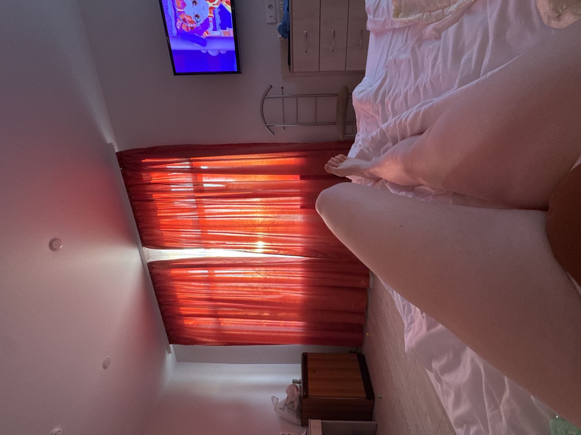 Here is a forensic description of the image:
**Location:** Appears to be inside a bedroom. The exact location in Saint Petersburg, Russia, cannot be determined from the image.
**Subjects:** Human legs and feet are visible in the bed.
**Objects:**
* Bed with white linens
* Red curtains
* Television displaying a cartoon
* White dresser
* Metal towel rack
* Nightstand
* Ceiling lights
**Environment:** The room is lit by daylight coming through the window and the television.