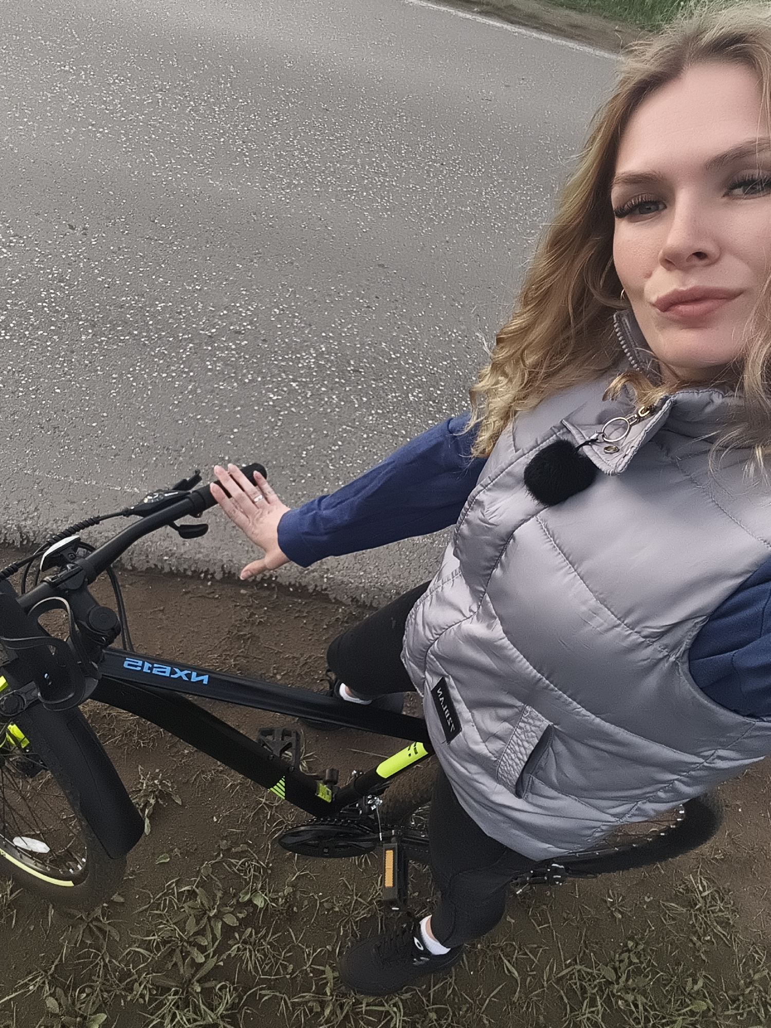 A woman is taking a selfie while on a bicycle. She is wearing a silver puffer vest over a blue long-sleeved shirt, and black leggings. She has blond hair and is wearing hoop earrings. The bicycle is black with blue lettering. The ground is a mix of pavement and dirt/grass. It is likely in Русский Вожой, Russia.