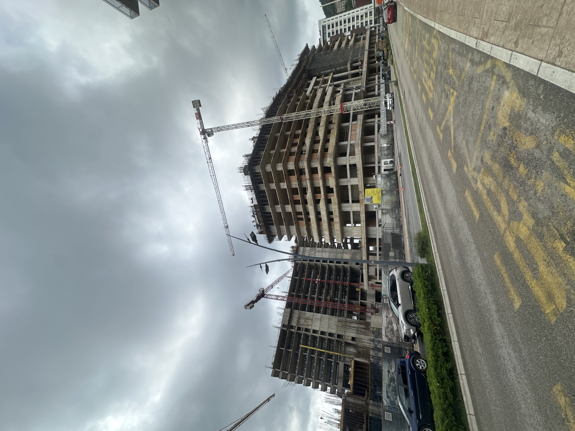 This image captures a large-scale construction site under an overcast sky, likely during daytime. The scene is dominated by multiple multi-story buildings in various stages of concrete frame construction, showcasing exposed rebar, concrete slabs, and scaffolding. Several large tower cranes, some white and some red, are positioned around and above the structures, indicating active development.
A paved road runs along the right side of the frame, parallel to the construction site. Faded yellow markings, which appear to spell out "PARKIRANJE" (parking), are visible on the asphalt. A strip of green bushes separates the road from the construction area. Several cars are parked along the road, including a silver SUV in the mid-ground and a dark blue sedan further back. Other vehicles, including a red car, are visible in the distance.
The sky is entirely covered by thick, gray clouds, suggesting a cloudy day without direct sunlight. In the background, glimpses of completed or nearly completed high-rise buildings can be seen. The overall impression is one of an expanding urban landscape with significant ongoing construction activity, consistent with an area experiencing development, such as Budva, Montenegro. No people are distinctly visible.
