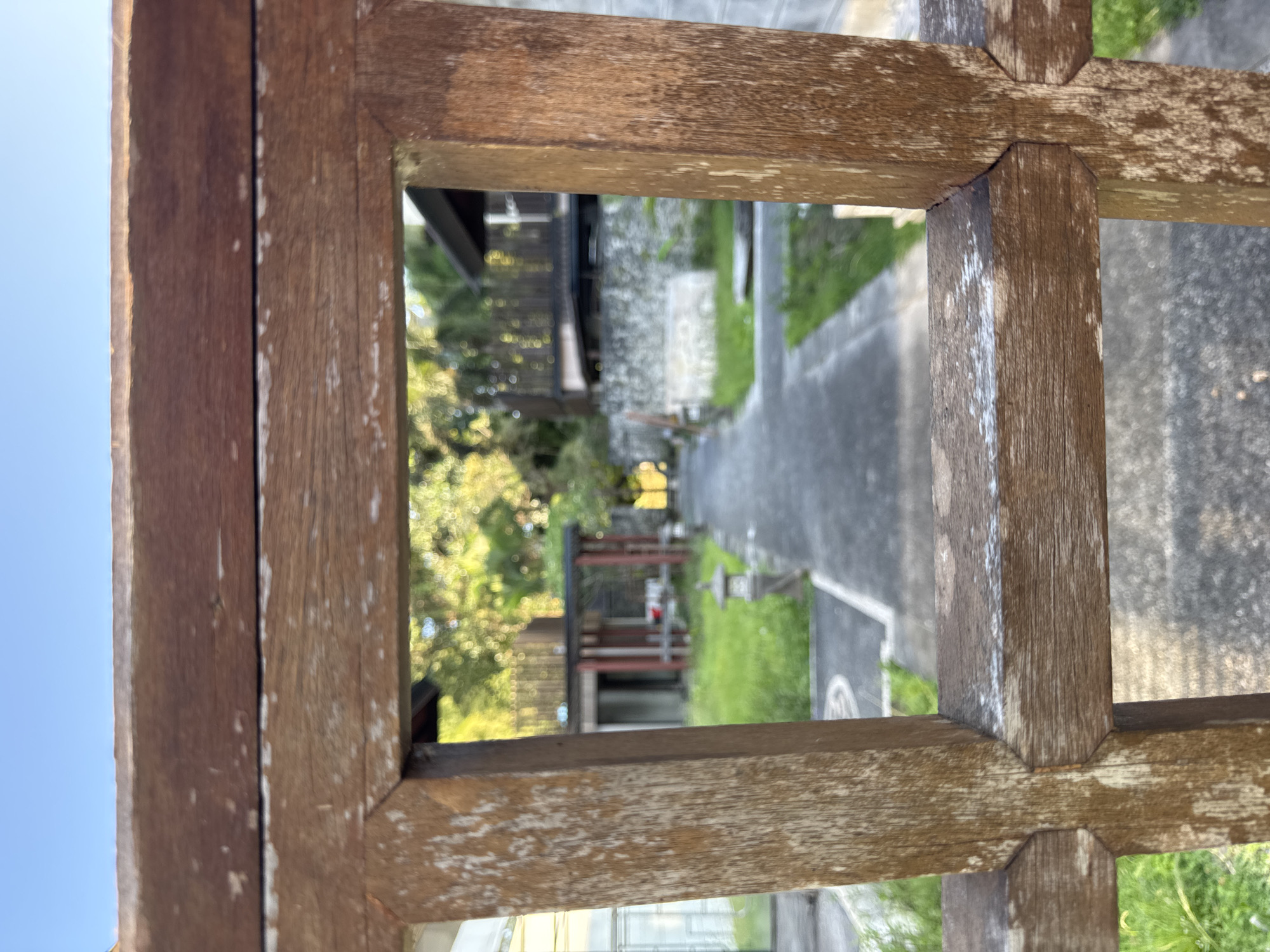 From an eye-level perspective through a weathered wooden frame, a pathway is visible in Kuta Selatan, Indonesia. The path, flanked by grassy areas, leads toward a building with a red roof. The sky is blue.
