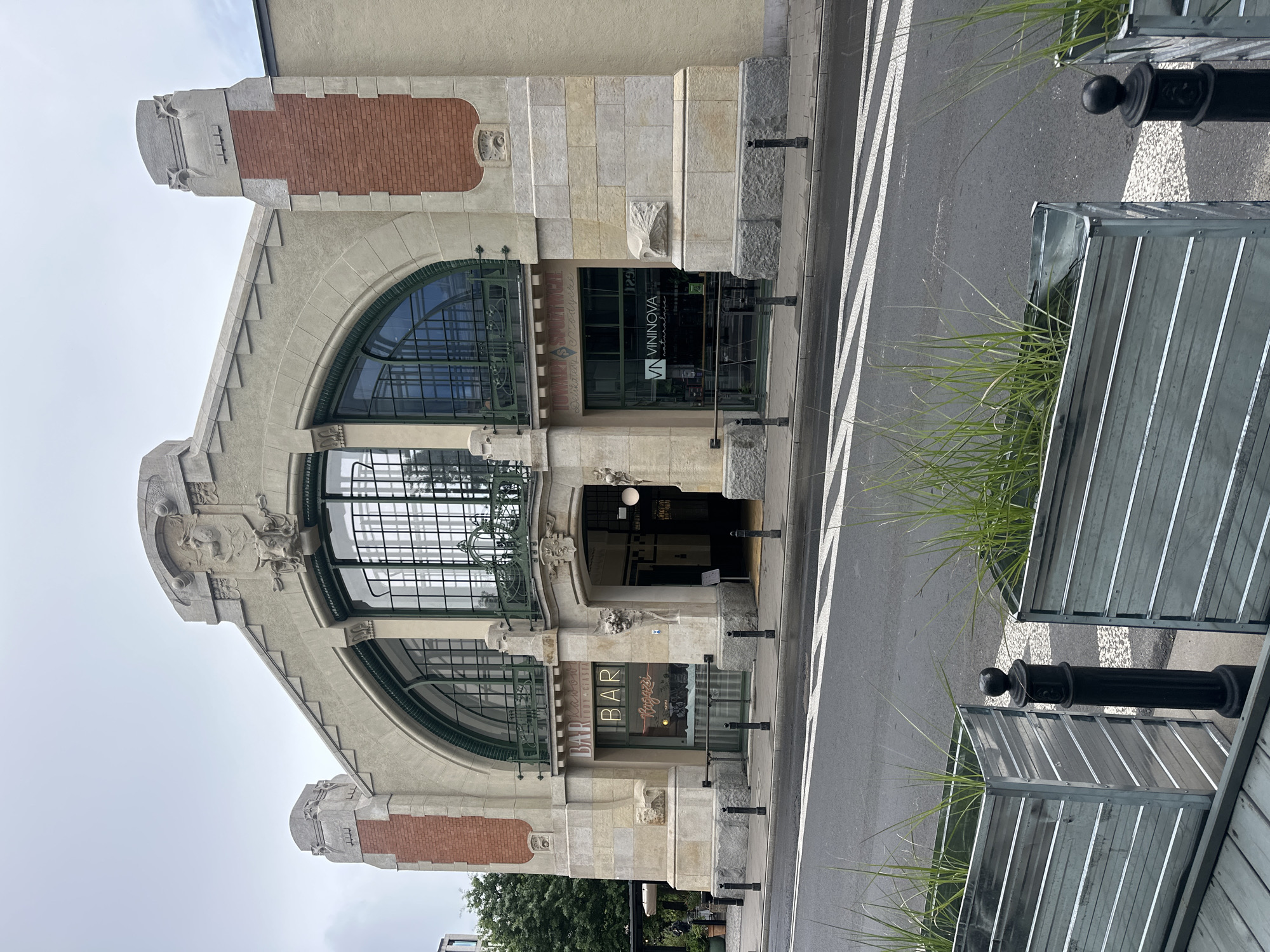 The image shows the entrance to a building with a large arched window and elaborate architectural details. There are signs for "BAR" and "VININOVA" visible on the storefronts. Planters with grass are in the foreground, and a street with a pedestrian crossing is in front of the building. This building is the former market hall of the Hala Mirowska, located in Warsaw, Poland. The style of architecture suggests it's an older, perhaps historical, building that has been repurposed. The sky is overcast, and the overall impression is one of a commercial street scene in a European city. No people are clearly visible in the image.