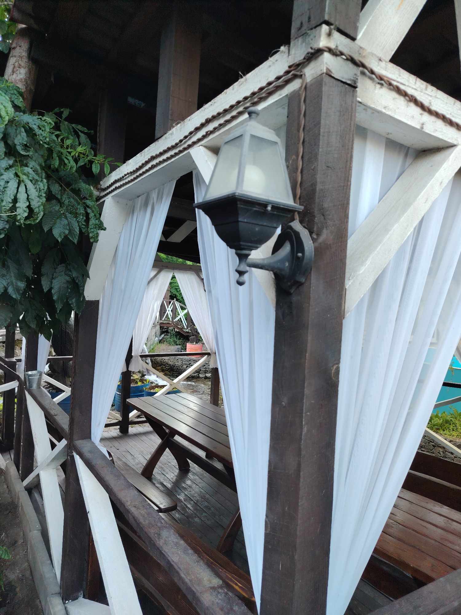 The image depicts an outdoor seating area, possibly a gazebo or covered patio, with white fabric draped along the sides. A wooden table and benches are visible. A dark-colored lantern is mounted on a wooden post. The setting appears to be a natural environment, possibly near water, with greenery and trees in the background. The architecture suggests a rustic or traditional style. The image does not contain any identifiable landmarks or clues that would place it specifically in Rostov-on-Don, Russia.