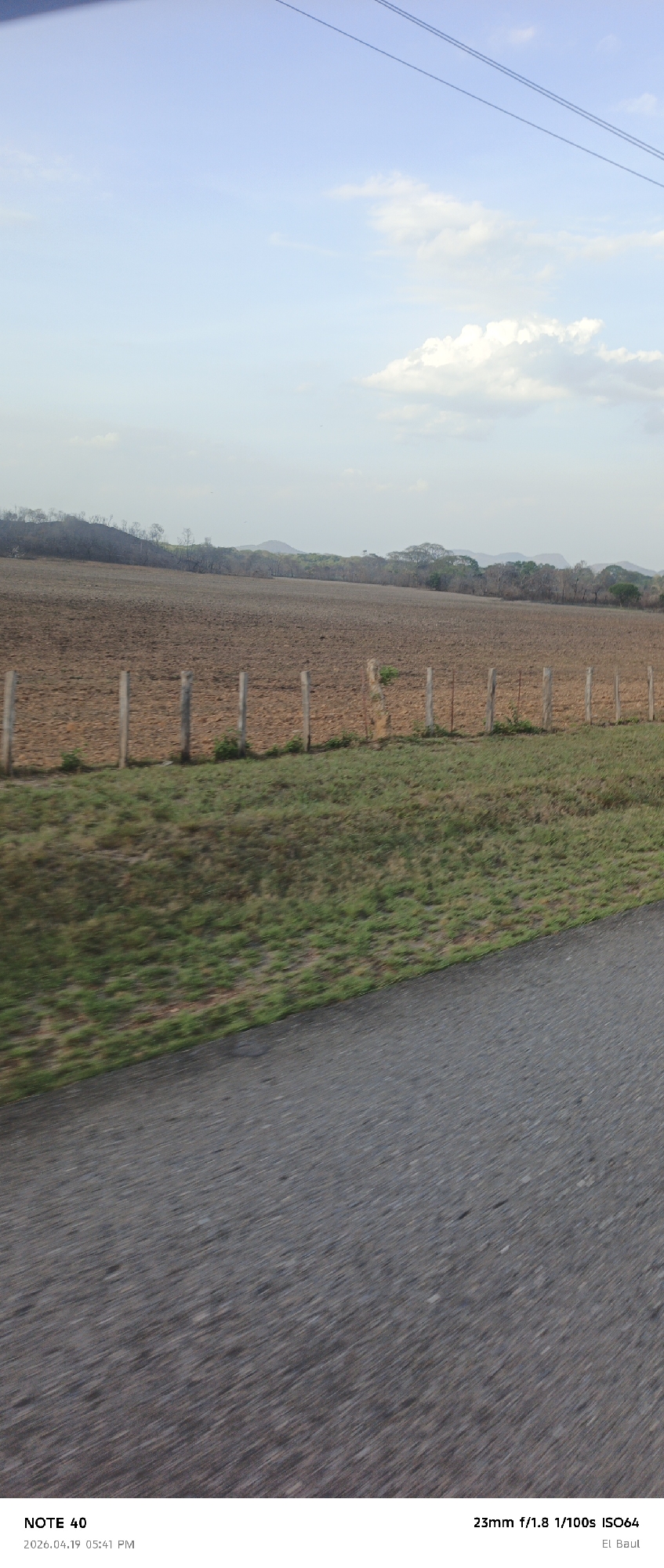 The image shows a rural scene with a focus on a plowed field bordered by a rudimentary wooden fence. Beyond the field, there are hints of distant hills and trees under a partly cloudy sky. In the foreground, the blurred surface suggests a road or path, indicating the image was likely taken from a moving vehicle. According to the OCR, the location is "El Baul", which is somewhere near La Cruz, Venezuela. The image captures a landscape view.