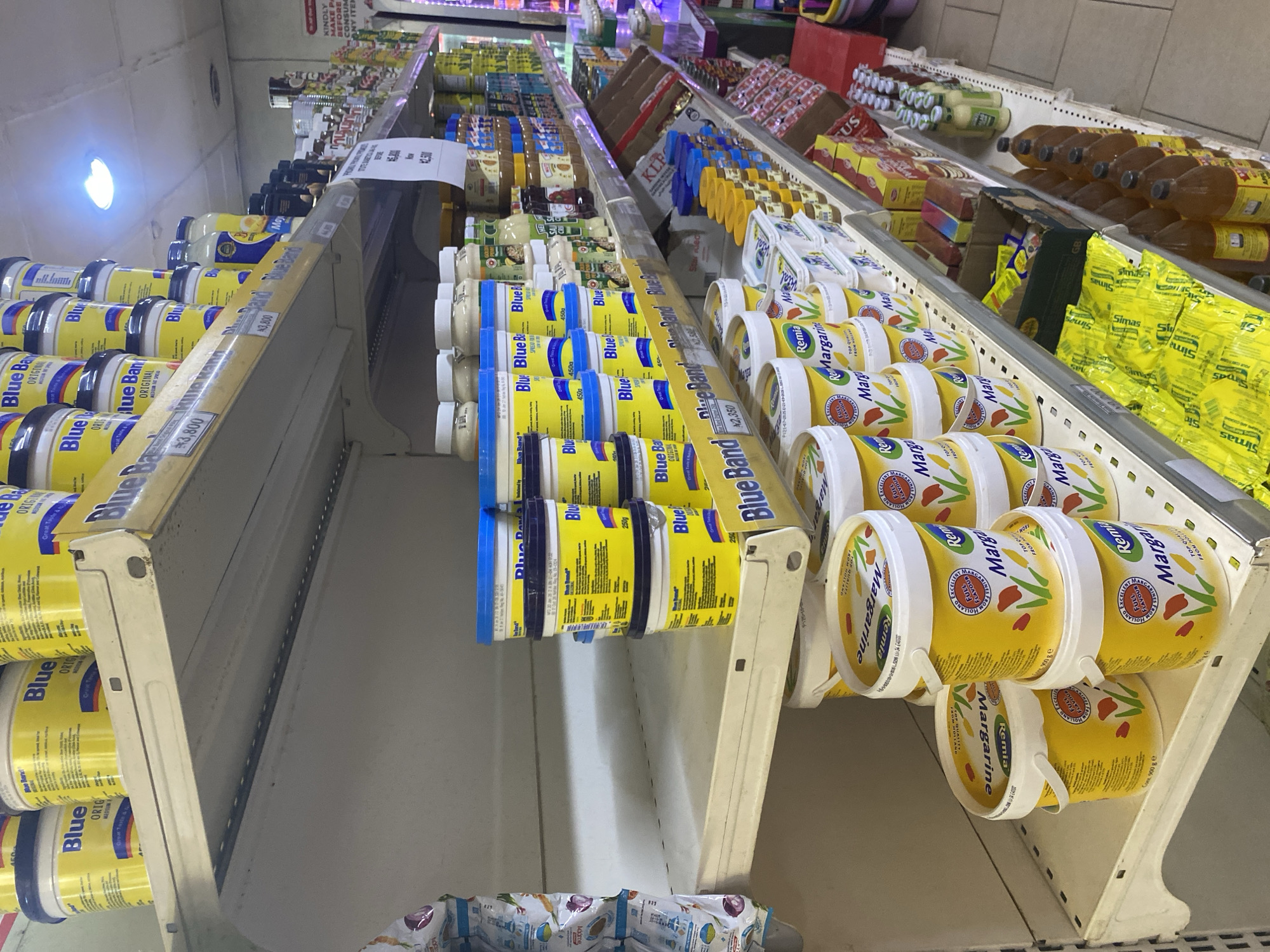 This is a forensic, matter-of-fact description of a supermarket shelf in Ikorodu, Nigeria. The image displays shelves stocked with various food products. The primary focus is on rows of yellow tubs labeled "Blue Band" and "Remia Margarine." The Blue Band tubs, in multiple sizes including 250g and 450g, are stacked prominently on the middle and upper shelves. These tubs are bright yellow with blue lids and feature images of the Blue Band logo and descriptive text. Several other brands and types of products are visible on the shelves, including what appear to be jars of spreads or sauces, canned goods, and packaged snacks. The shelving units are made of metal, with multiple tiers visible. In the background, other shelves and aisles of the supermarket are partially visible, with some lighting fixtures on the ceiling. The overall environment suggests a well-stocked retail setting. No people are visible in the image.