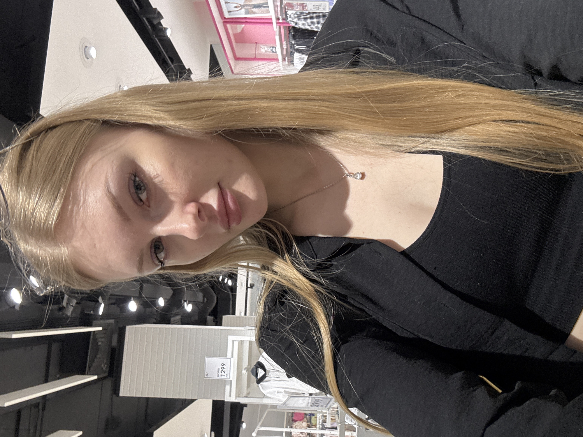 A young woman with long blonde hair is posing for a selfie in a store. She is wearing a black top and a silver necklace. In the background are clothing racks with white shirts and price tags. The price tag visible reads "1299" in Russian currency. The store appears to be brightly lit with fluorescent lights. No specific evidence indicates the exact location as Perm, Russia. The context suggests the woman is shopping.