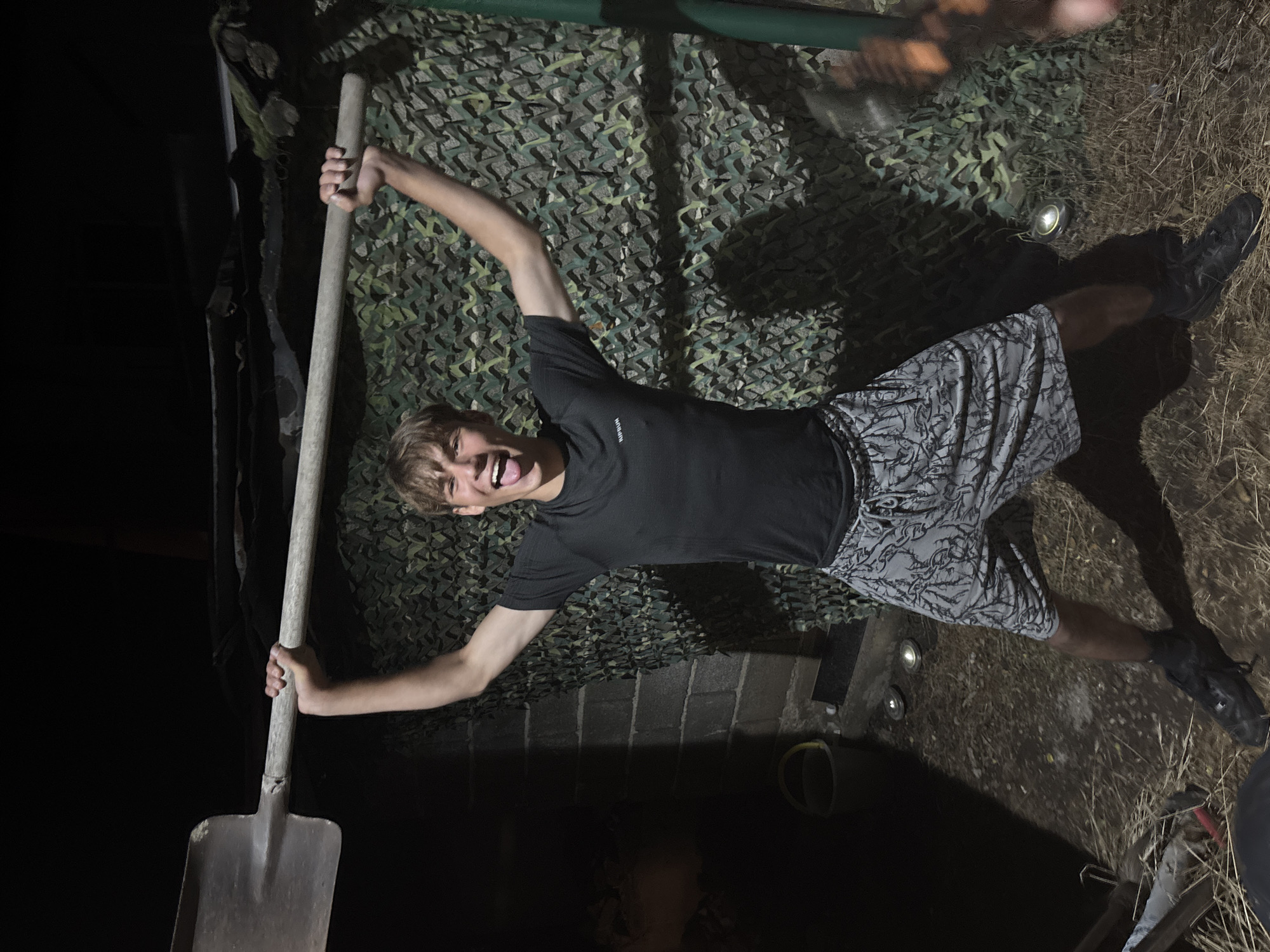 A young male, appearing to be in his late teens, stands outdoors at night, holding a large shovel above his head with both hands. He is wearing a black t-shirt and grey patterned shorts, along with black athletic shoes and dark socks. His tongue is sticking out, and he has a playful expression. The background features a brick wall on the left and a camouflage net draped over a structure on the right. The ground is covered with dry grass and dirt. There are a few small lights visible on the ground, and a yellow bucket is positioned near the brick wall. The overall lighting suggests it is nighttime, with some artificial light sources illuminating the scene.
