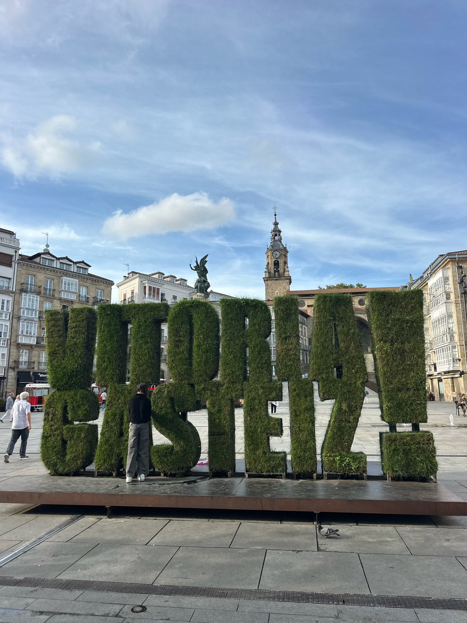 The image displays a large, sculpted sign made of lush green grass, spelling out "VITORIA GASTEIZ." The sign is situated in a paved public square with buildings and a clock tower visible in the background. Two individuals are standing near the sign; one is a person with dark hair wearing a black top and grey pants, facing away from the camera and looking at the grass sculpture. The other person, to the left, is wearing a white shirt and dark pants, also facing away from the camera. The sky above is blue with scattered white clouds. In the distance, part of a building with multiple windows and balconies is visible. The overall scene suggests an outdoor urban environment, likely a plaza or a prominent public space within the city of Vitoria-Gasteiz, Spain.