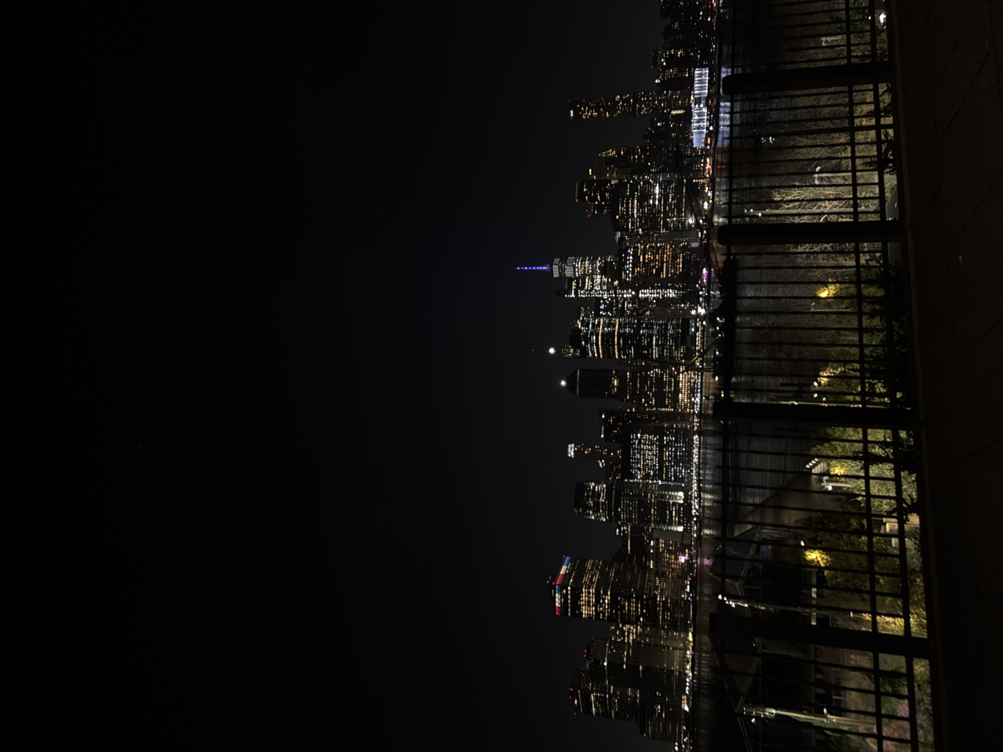 The image shows a night view of the New York City skyline from a balcony or elevated position. The city is brightly lit with numerous skyscrapers. To the right, a portion of a fence or railing is visible, suggesting the viewpoint is from a building or terrace. The sky is dark, contrasting with the illuminated buildings.