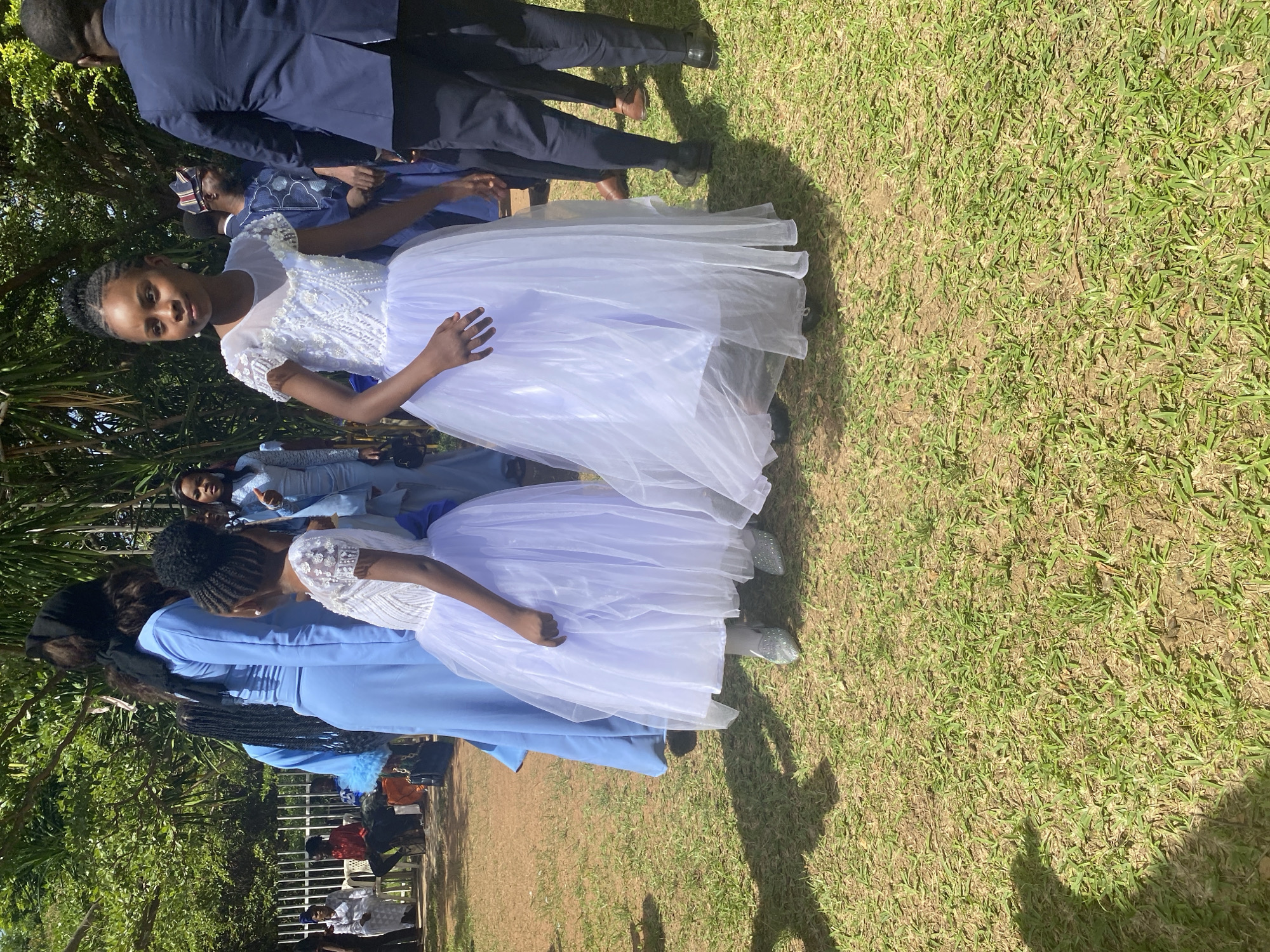 The image captures a scene in Bukuru, Nigeria, featuring a group of people on a grassy area. Two young girls are prominently displayed, both wearing white dresses with decorative tops and blue sashes. They appear to be part of a formal event. Several adults are also present, some dressed in blue formal attire and others in dark suits. The background includes trees and glimpses of other attendees seated. The light suggests it is daytime, and shadows are cast on the grass.
