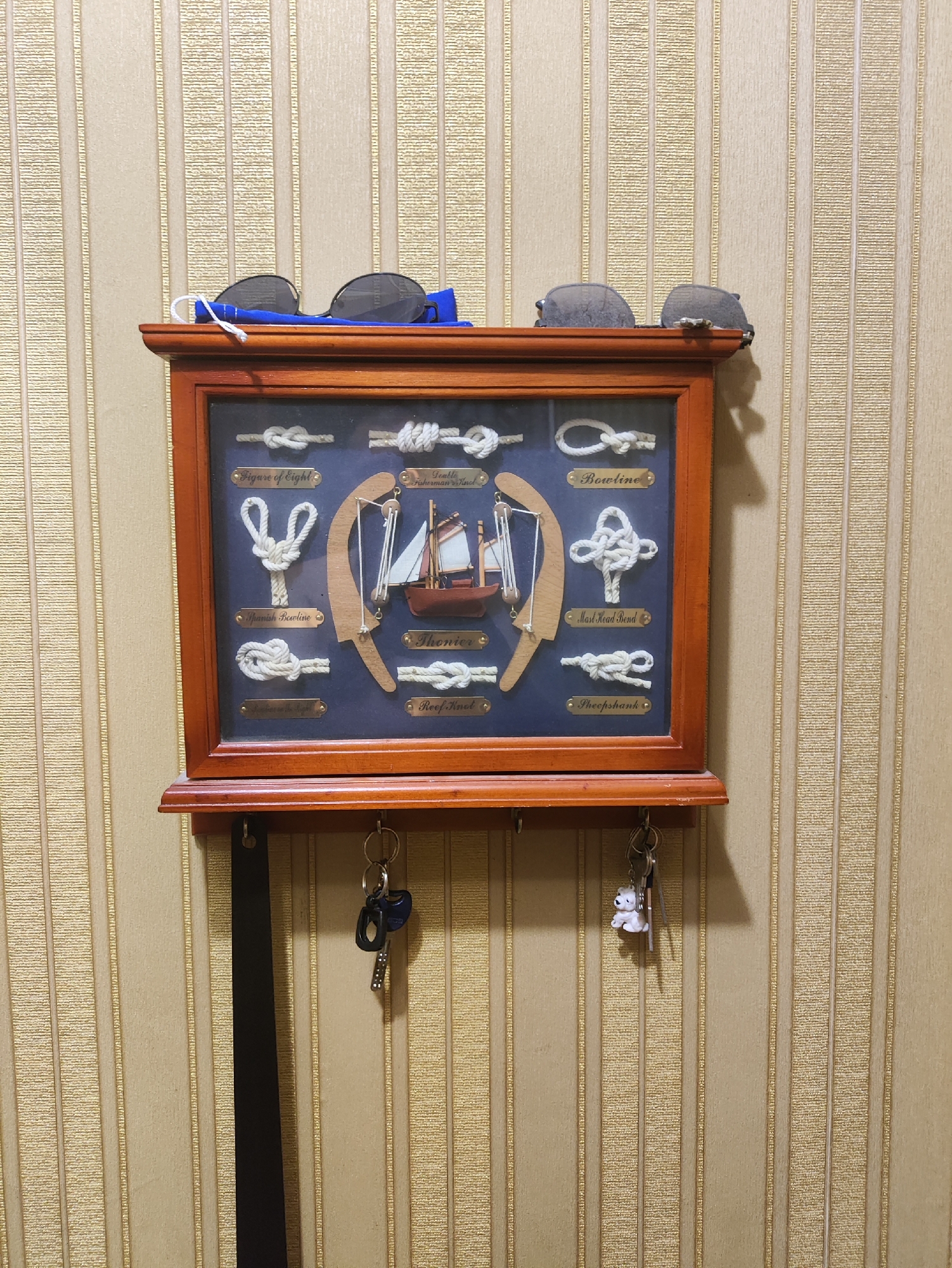 A wooden key holder with a glass-covered display of nautical knots hangs on a beige, vertically striped wallpaper. Several pairs of sunglasses rest on the top edge of the key holder. Below the display, multiple keys, including one with a small white dog charm, hang from hooks. The scene appears to be an interior domestic setting. There is no information to indicate the location in Kharkiv, Ukraine.