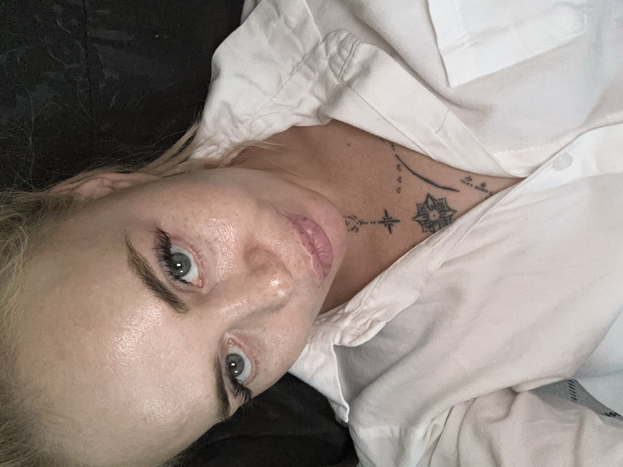 A close-up, overhead shot shows a blonde woman with light blue eyes looking down and slightly to the left. She is wearing a white collared shirt, partially unbuttoned, revealing a tattoo on her chest and neck area. The tattoo appears to be intricate with geometric shapes and floral designs. Her skin is fair with visible freckles, and she has on mascara and light-colored lipstick. Her hair is pulled back, with some strands falling loose around her face. The background is dark and indistinct, suggesting an indoor setting.