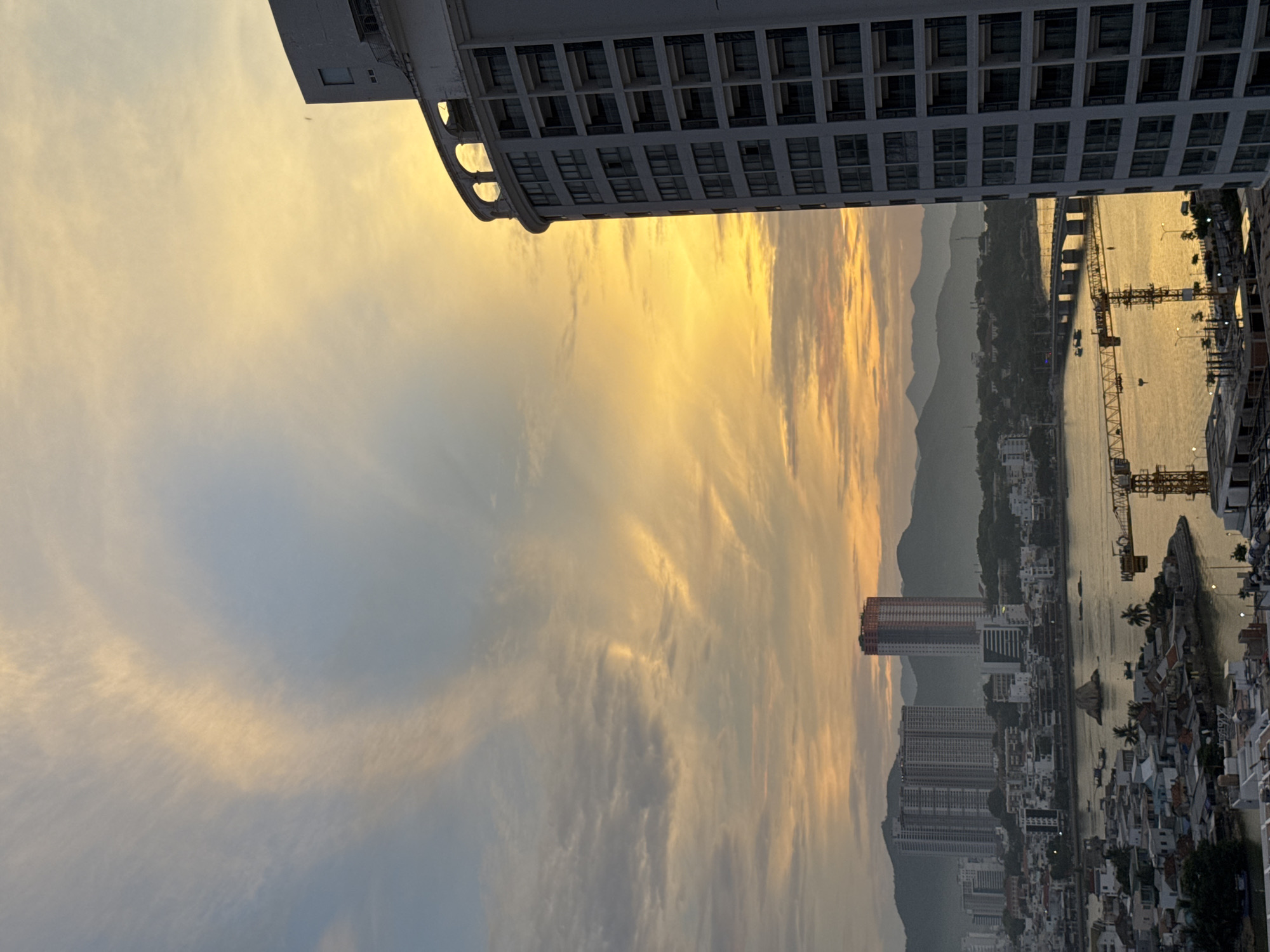 The image depicts a coastal cityscape of Nha Trang, Vietnam, during a vibrant sunset. The sky is filled with a dramatic display of golden and orange clouds, transitioning into a pale blue hue towards the top. In the foreground, the side of a modern, multi-story white building with numerous windows is prominently featured on the right, its curved architectural elements visible against the sky.
Beyond the building, the city stretches out towards a range of hazy, mountainous terrain in the distance. A body of water, likely a river or bay, separates the city from the mountains, reflecting the warm colors of the sunset. Several smaller boats are scattered across the water. A bridge spans across the water, connecting different parts of the city. In the middle ground, a cluster of buildings, including tall apartment complexes and a distinctive red and white skyscraper under construction, are visible. Yellow construction cranes are also present, indicating ongoing development. The lower parts of the city show a denser arrangement of smaller buildings. The overall scene is one of urban life set against a beautiful natural phenomenon.