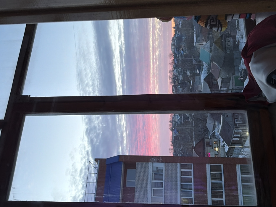 The image depicts a winter residential cityscape in Izhevsk, Russia, viewed through a dark brown wooden window frame. The window panes are slightly smudged and exhibit minor reflections. On the windowsill, folded red and white fabric, possibly clothing, and a small, light-colored object are visible in the lower right.
Outside the window, to the left, stands a multi-story red brick building with visible light-colored (white/grey) balconies. This building is topped by a distinctive blue mansard-style roof with a flat, dark-railed section. Further into the midground, a densely packed area of low-rise residential houses stretches across the landscape. These houses vary in size and roof color, with many appearing to be older structures, some featuring green or light blue roofs. The ground and the roofs of these houses are covered in snow. Sparse, bare trees are visible between the houses and along the distant horizon. One house in the mid-distance displays a bright pink light emanating from an interior window.
The sky occupies the upper portion of the view, presenting a dramatic horizontal gradient. It transitions from vibrant pink and reddish hues at the lower horizon, characteristic of sunrise or sunset, to lighter shades of blue and greyish-white towards the zenith. The sky is populated by elongated, stratified cloud formations.
The scene captures a residential sector of Izhevsk, characterized by the juxtaposition of a modern multi-story building and an older, lower-density private housing area, all under a winter sky at either dawn or dusk.